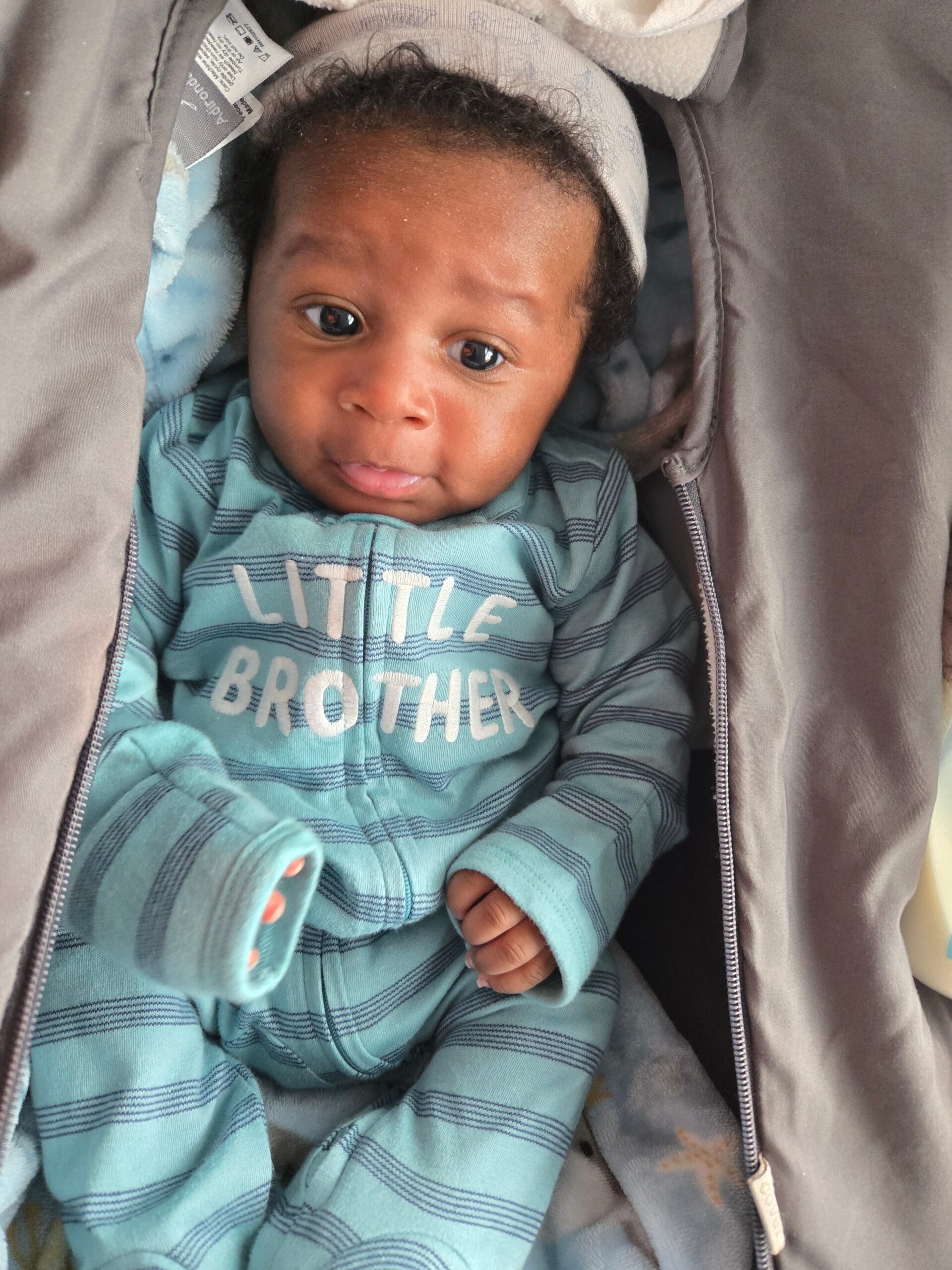 Close-up of a baby in a stroller wearing a teal striped onesie that says 'LITTLE BROTHER'.