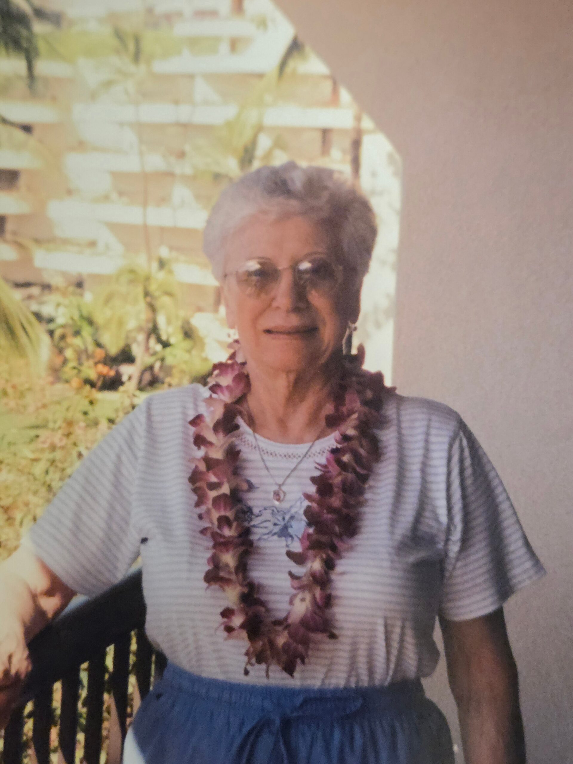 Older woman with short gray hair and sunglasses, wearing a lei and striped shirt, standing outdoors in a garden.