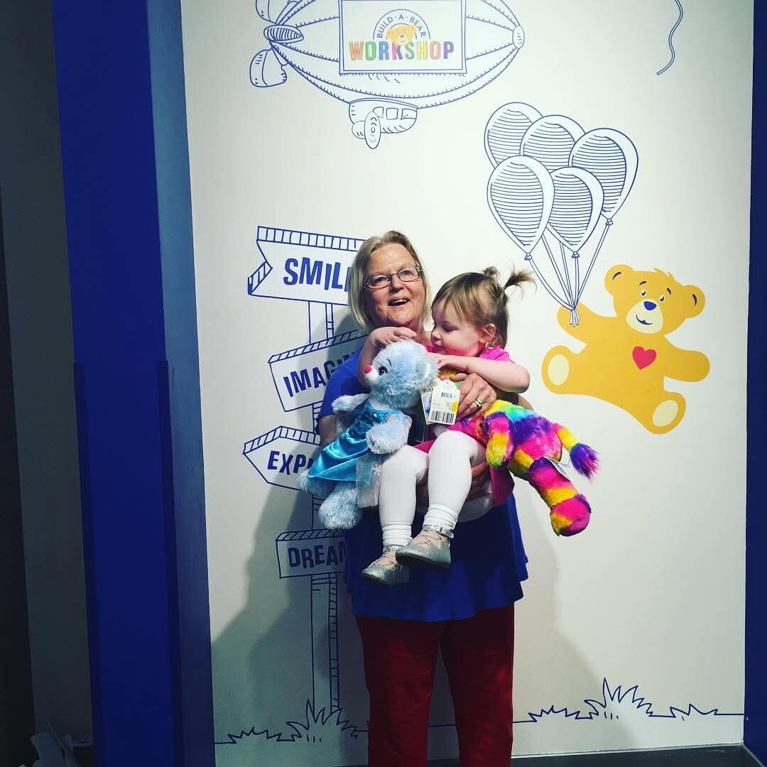 An older woman holding a young girl who clutches stuffed animals, posing in front of a playful mural with balloons, a teddy bear, and a 'SMILE' sign.