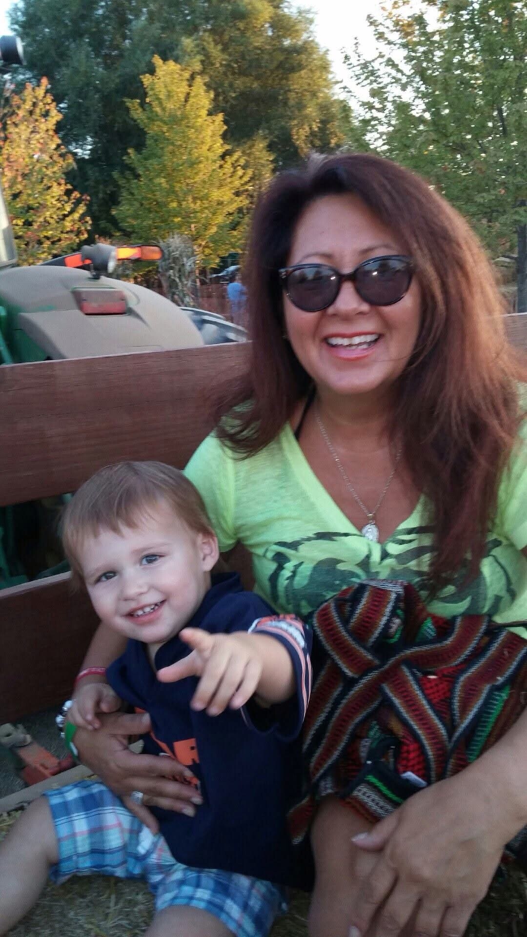 Smiling woman wearing sunglasses sits outdoors on a wooden bench with a young boy on her lap in a park setting.