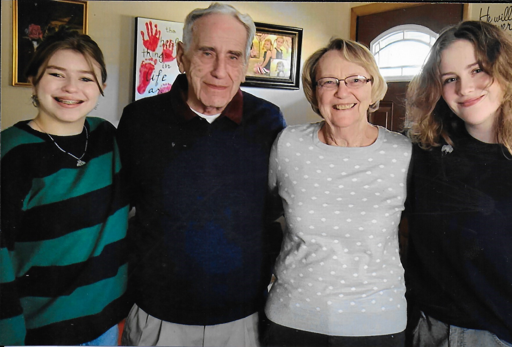 Four people stand together indoors, smiling for a group photo in a living room with framed art in the background.