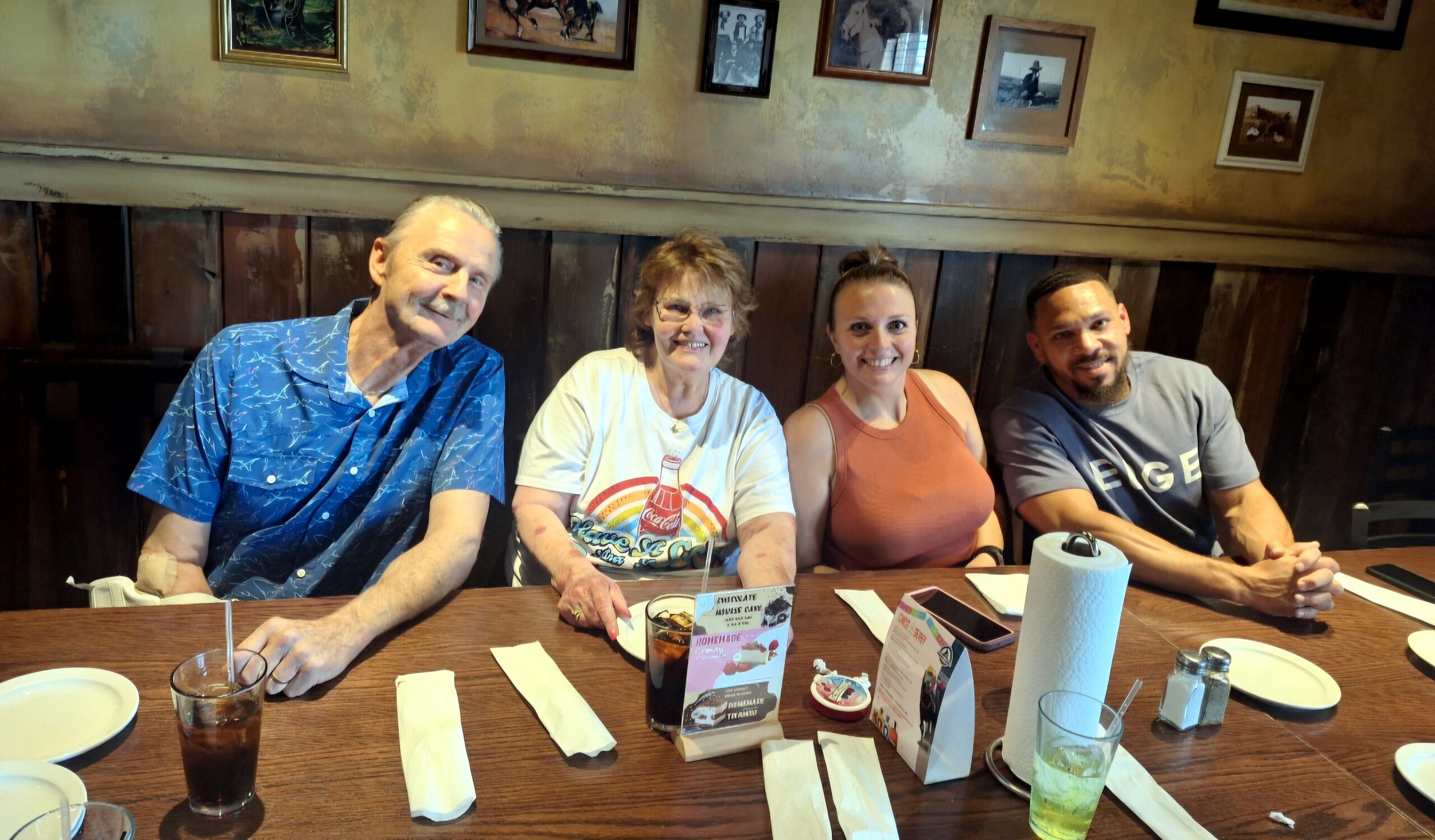 Four adults seated at a wooden restaurant table, smiling at the camera with drinks, napkins, and menus in front.