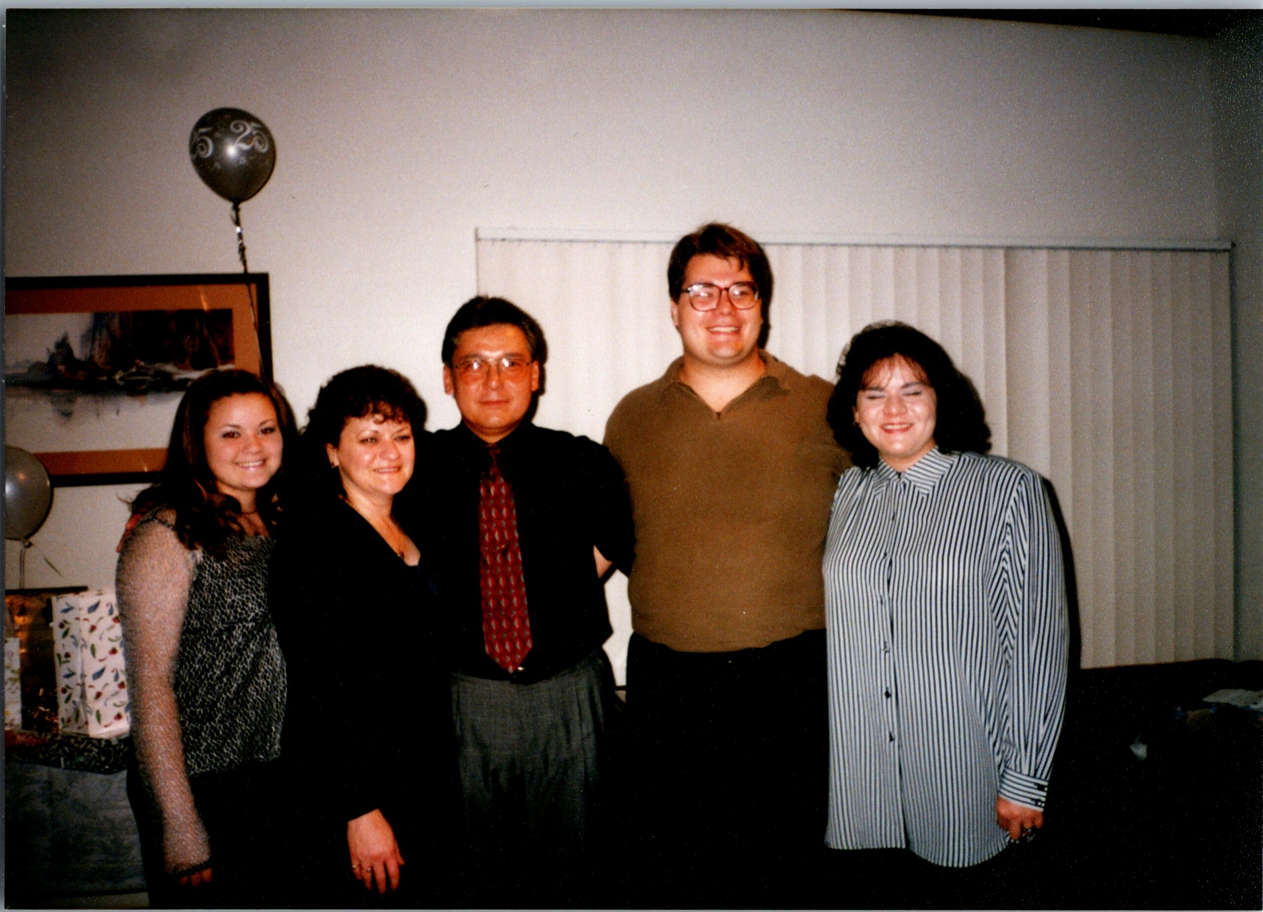 Five adults pose together indoors at a party, with a balloon visible in the background and everyone smiling.
