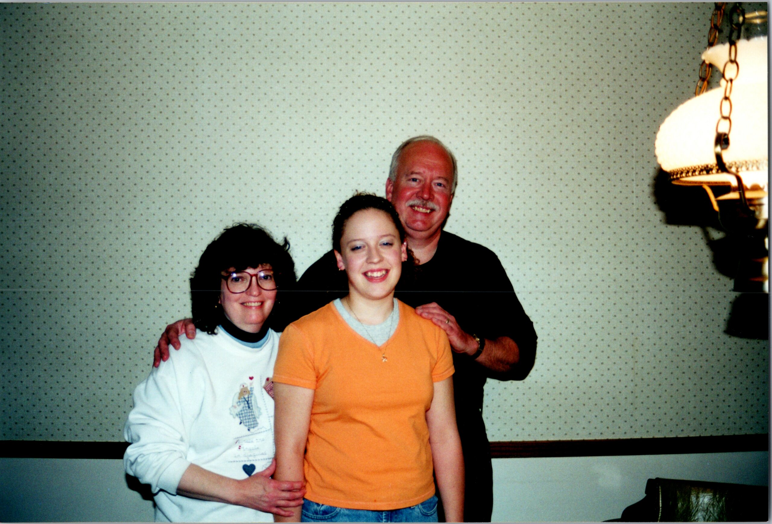 Three people pose together and smile for a casual photo in a room with dotted wallpaper (woman with glasses on left, girl in orange shirt front, man behind).