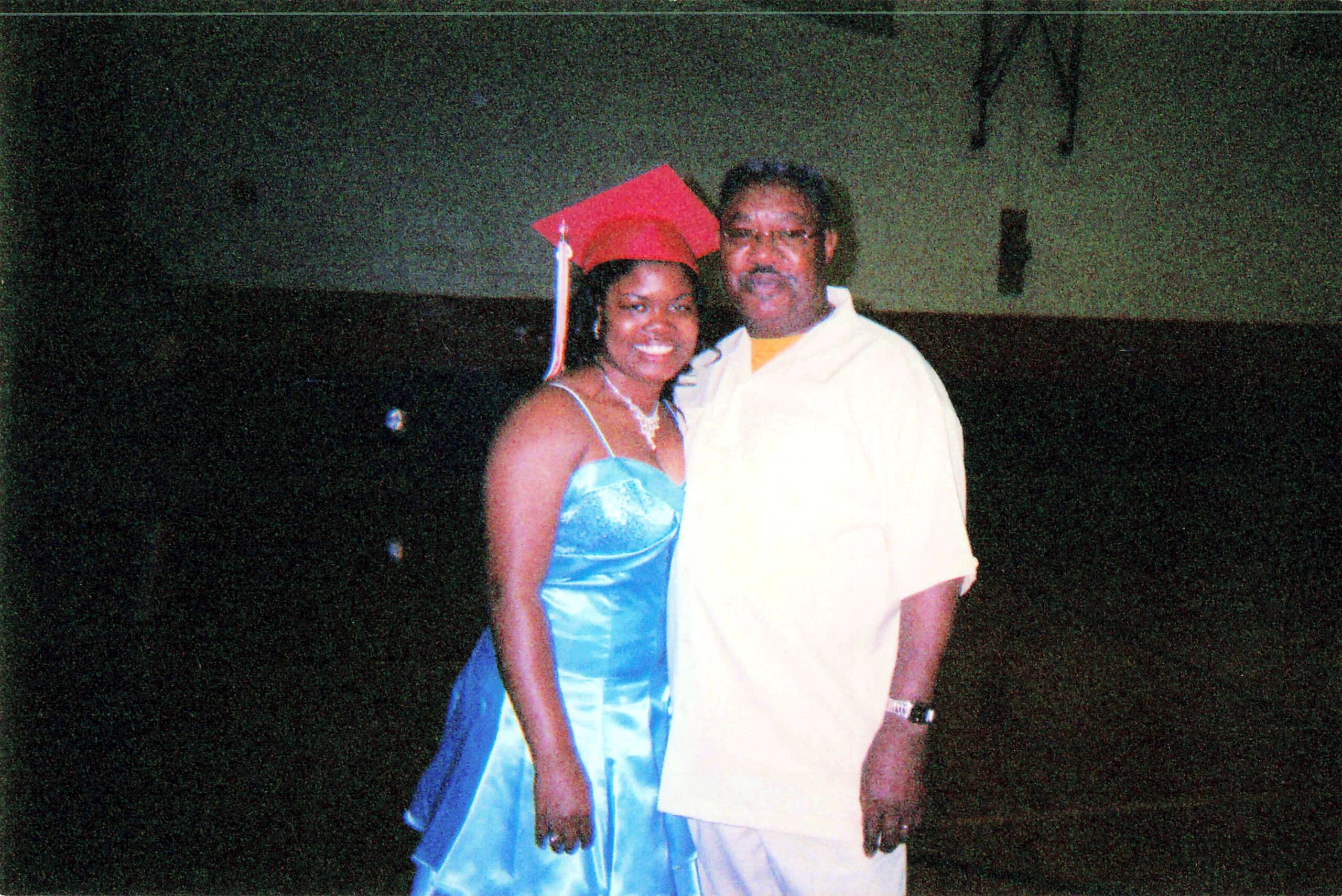 Smiling couple at a graduation: woman in a blue gown and red mortarboard beside a man in a light shirt during nighttime outdoors.
