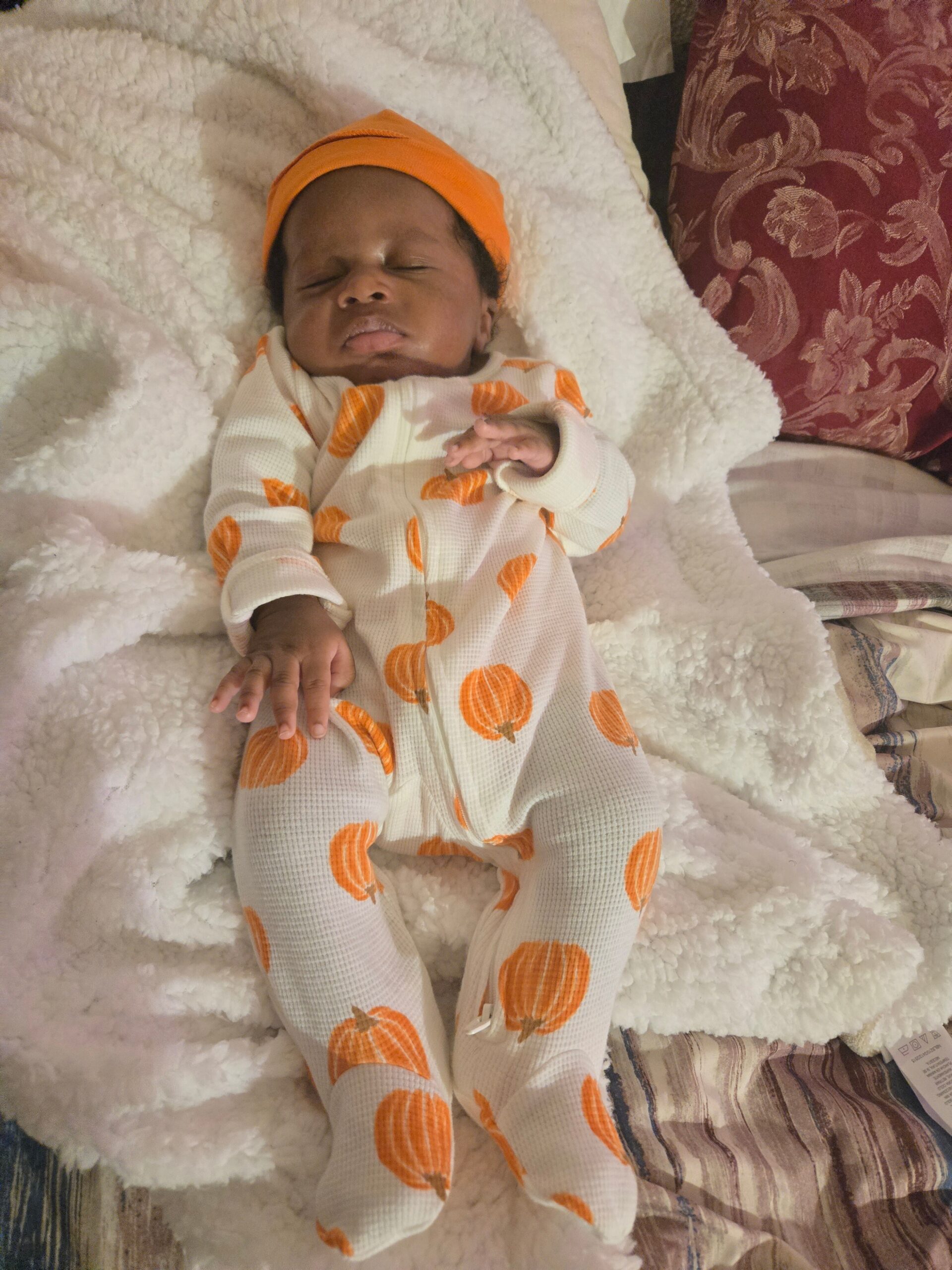 Newborn baby sleeping on a white fluffy blanket wearing an orange hat and cream onesie with orange pumpkins pattern.