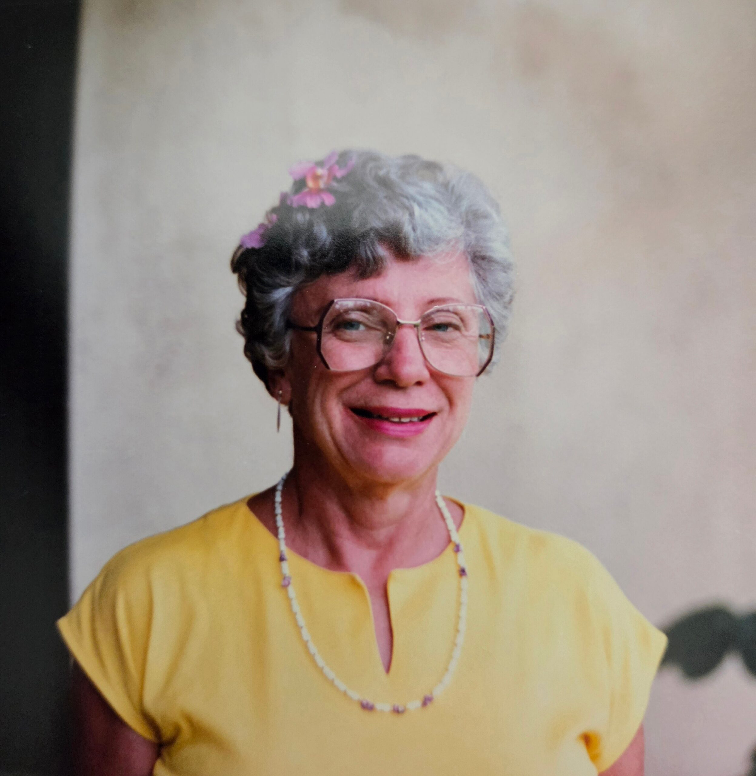 Smiling elderly woman with short gray hair and glasses, pink flower hair accessory, wearing a yellow blouse and pearl necklace.