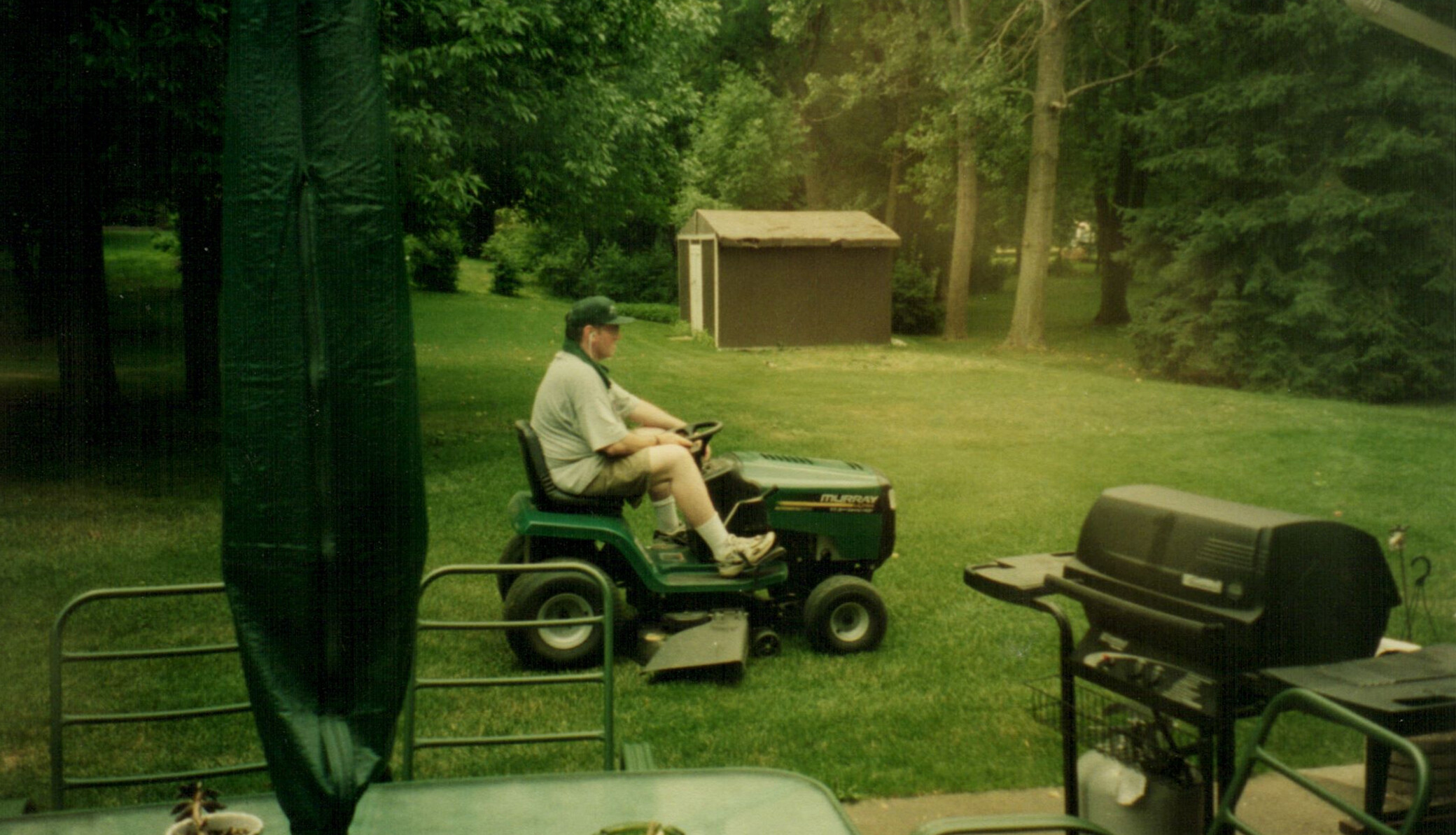 Man wearing a cap rides a green riding lawn mower across a grassy yard with trees and a brown shed in the background.