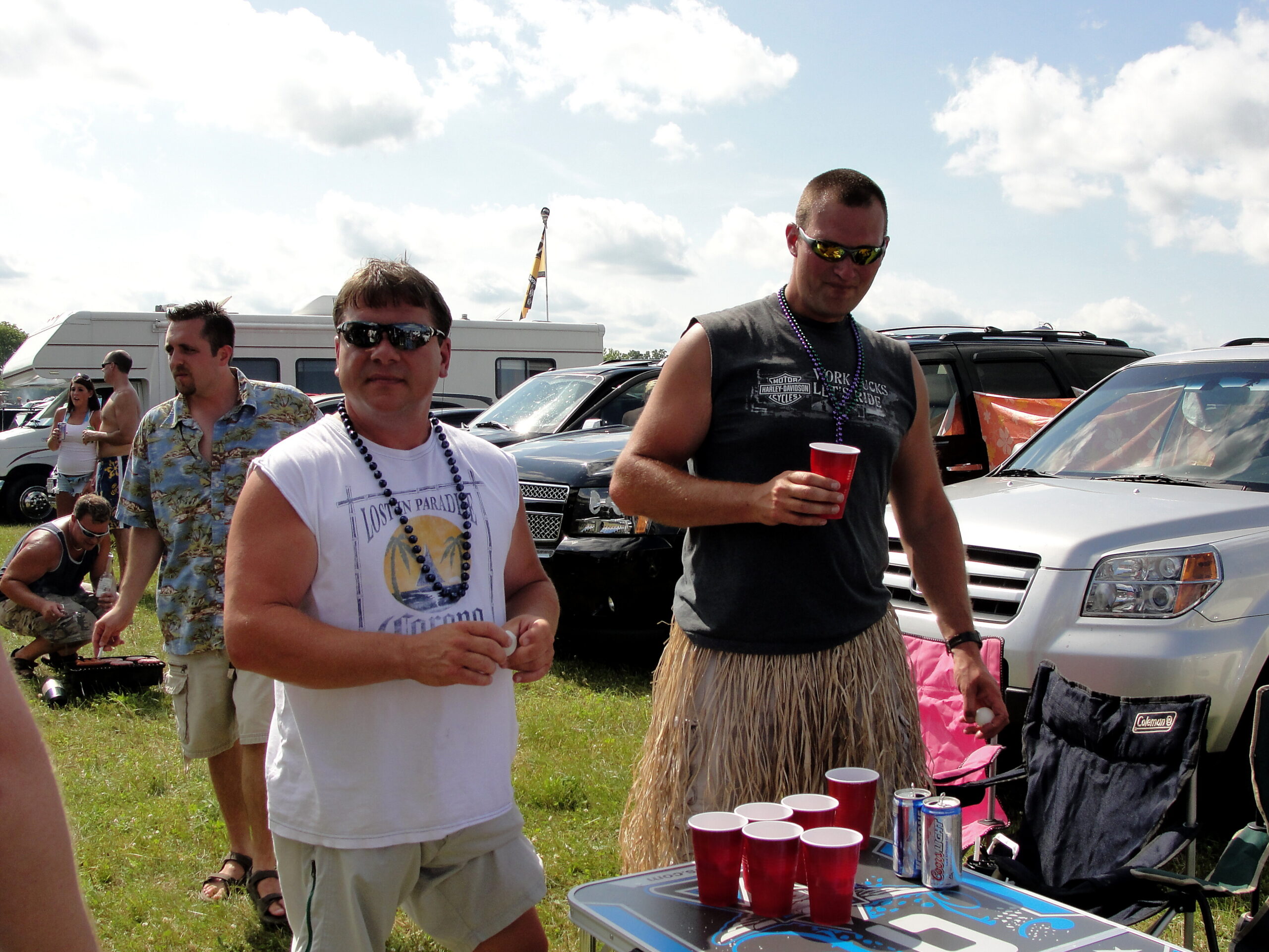 Group of adults at an outdoor tailgate party with red cups and snacks; man in white shirt foreground, man in black sleeveless shirt with sunglasses and bead necklace nearby.