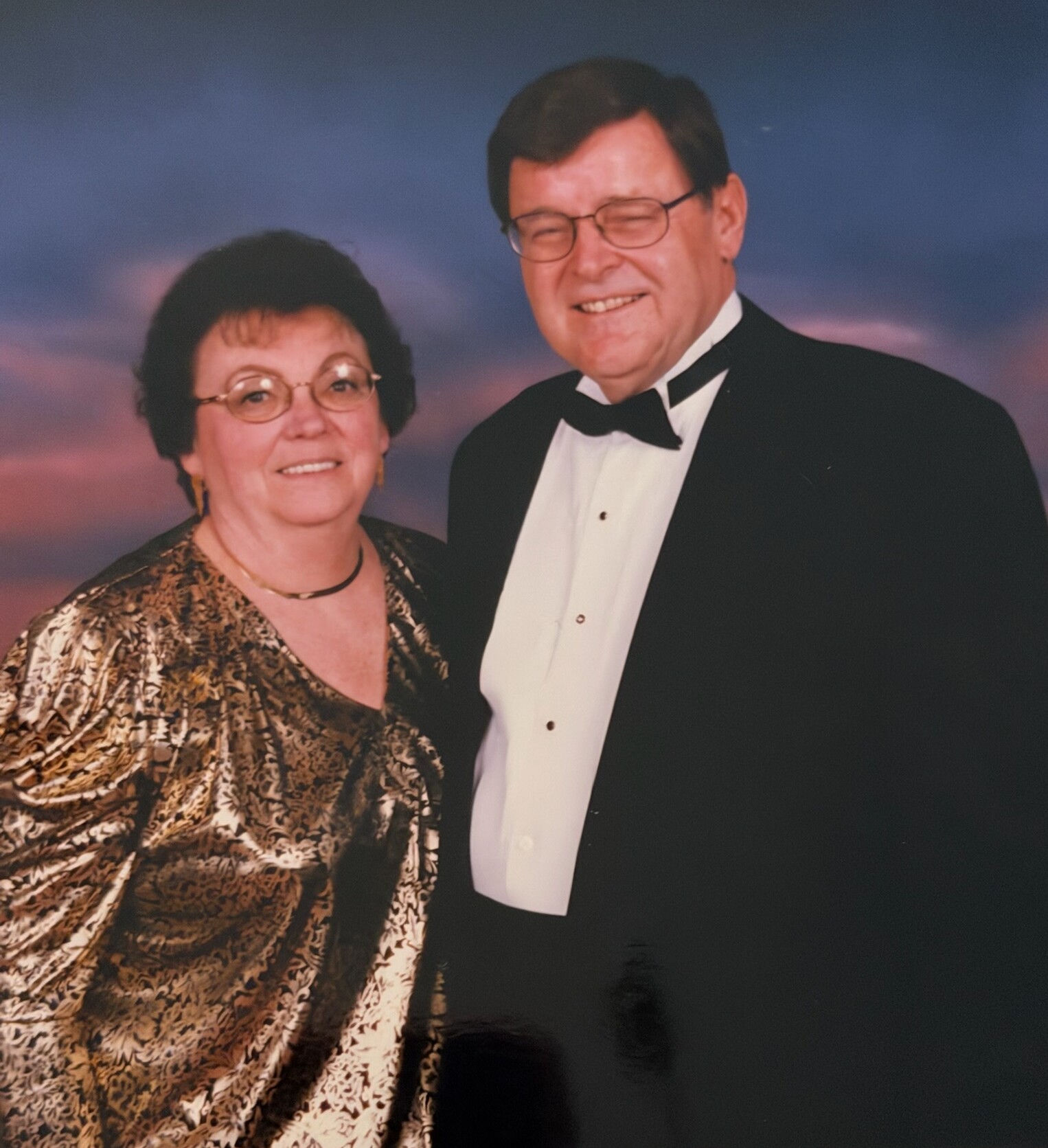 Smiling couple in formal attire posing together against a sunset backdrop. The woman wears a gold patterned dress; the man wears a tuxedo with bow tie.