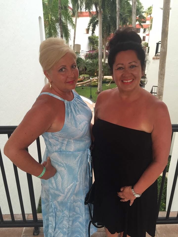 Two women stand on a balcony smiling; left is blonde in a light blue halter dress, right is brunette in a black strapless dress, with palm trees behind them.