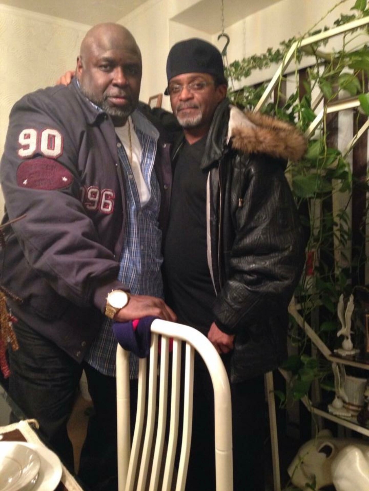 Two men pose closely indoors; one wears a gray varsity jacket with 90/96 patches, the other a black cap and fur-trimmed jacket, with plants in the background.