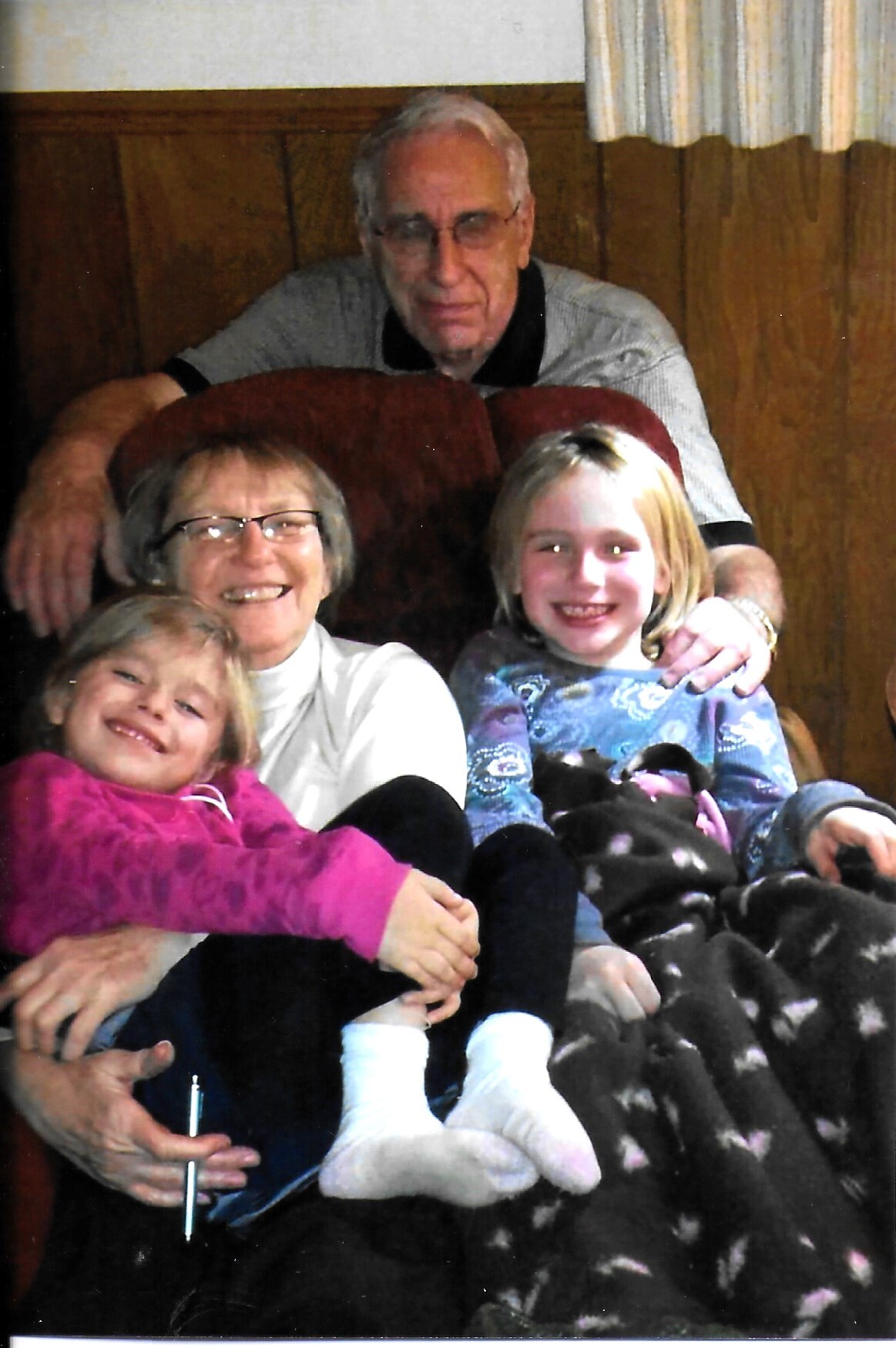 Grandfather standing behind a burgundy couch with a smiling mother and two young girls seated in front in a cozy room.