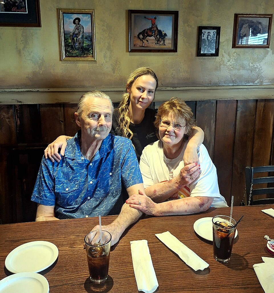 A woman stands behind an older couple with arms around them at a wooden restaurant booth, smiling at the camera, drinks on the table.