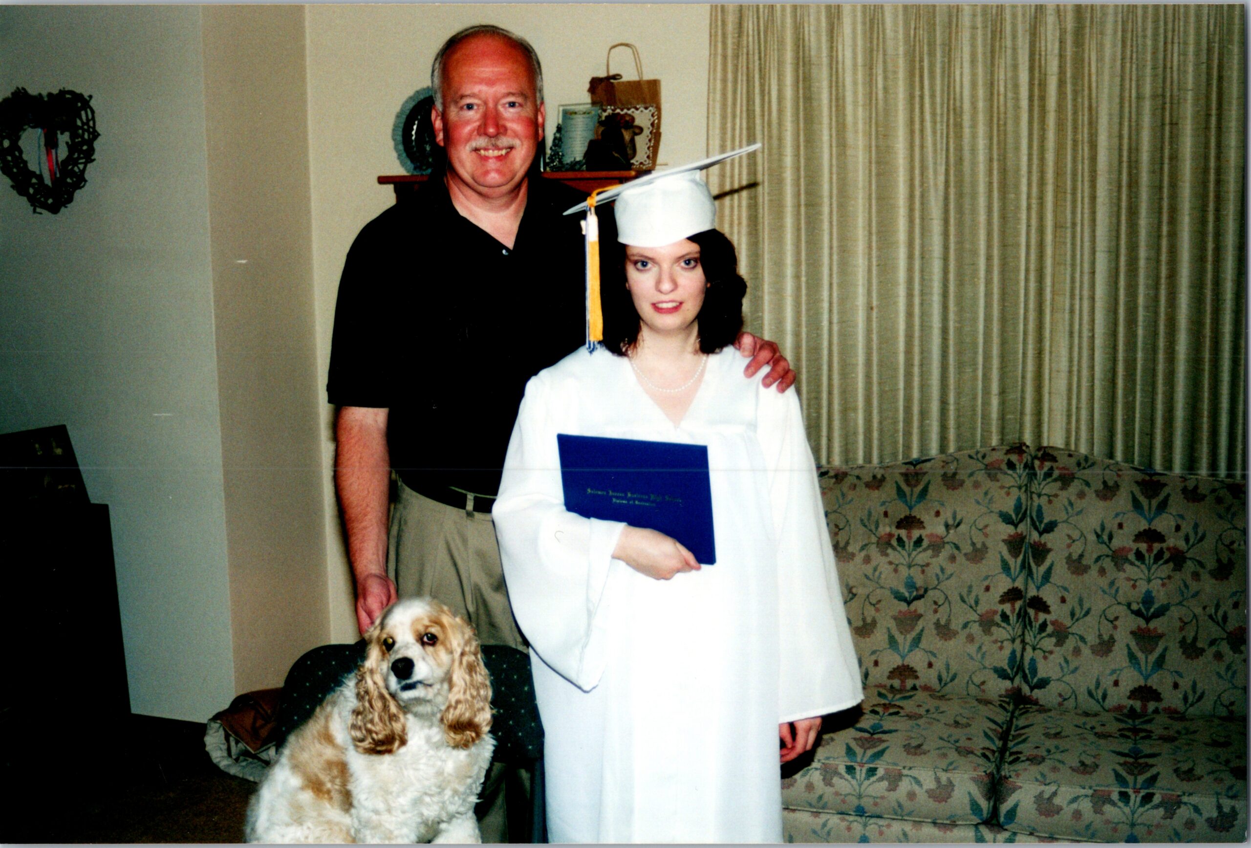 Older man stands beside a young graduate woman in a white cap and gown, holding a blue diploma; a dog sits nearby in a living room.