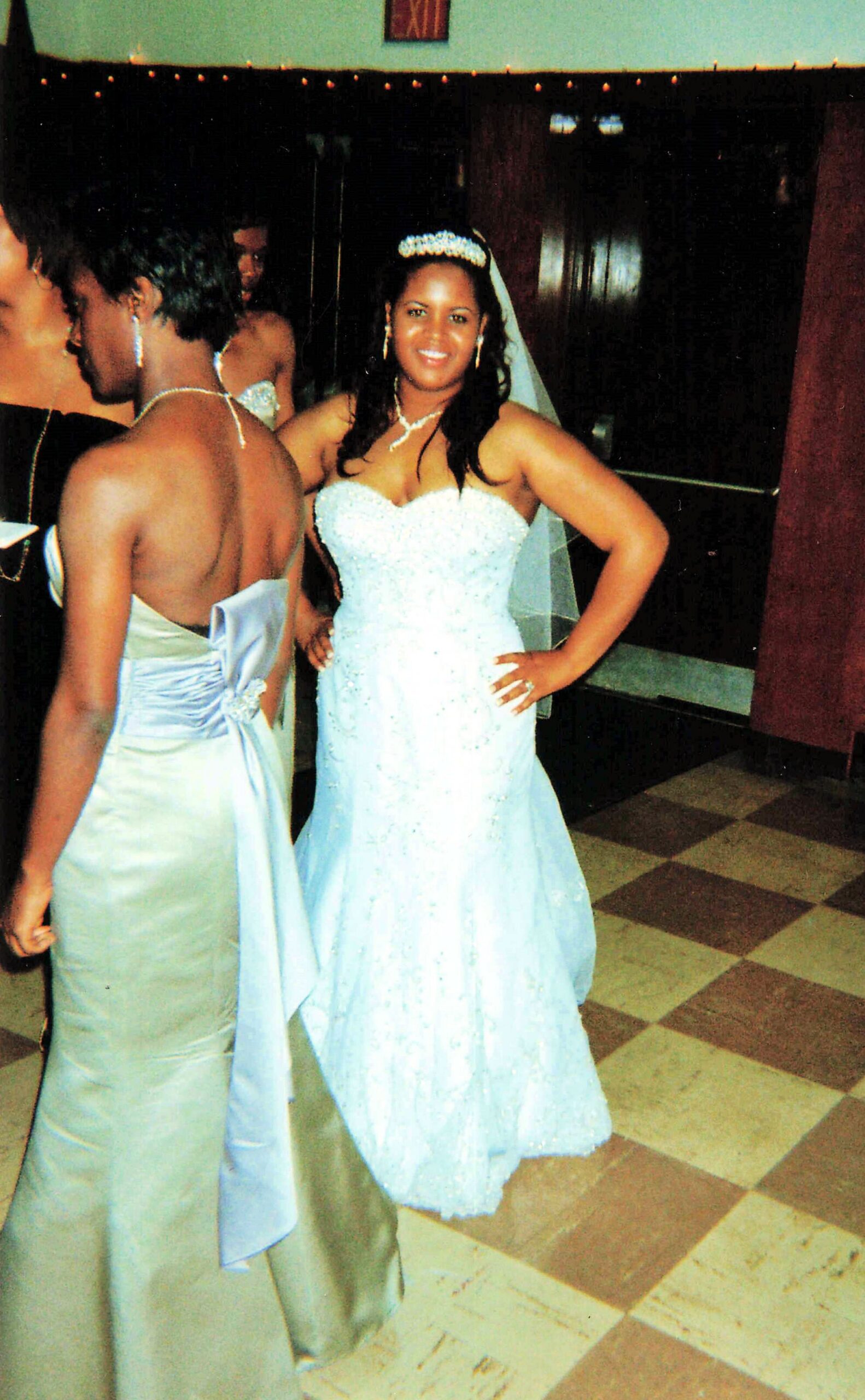 Bride in a sparkling light-blue strapless gown with a tiara and veil, posing with hand on hip at a formal event; other guests in gowns in the background.