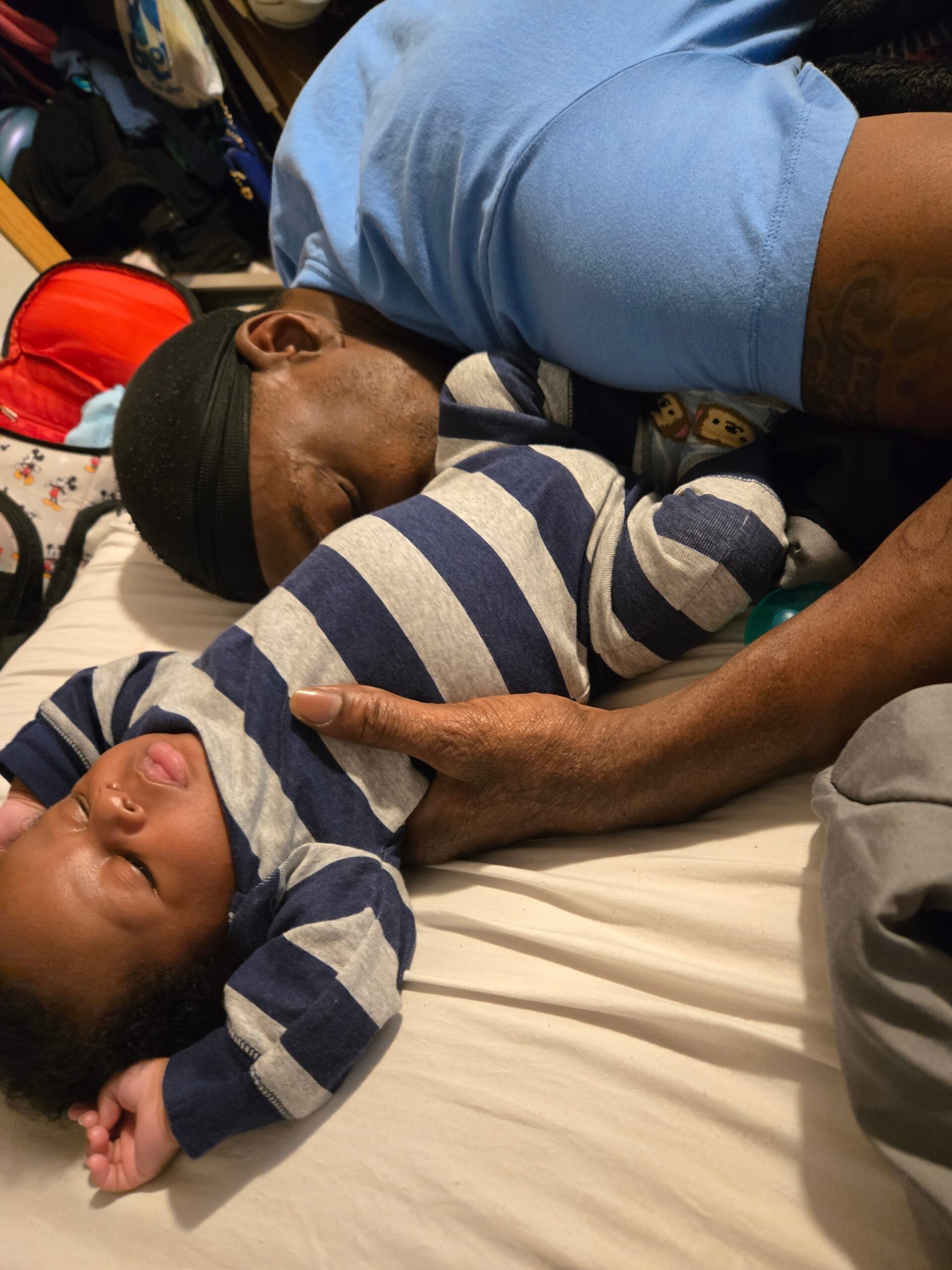 Parent lying beside a sleeping baby in a blue outfit on a bed, with the caregiver’s arm resting nearby