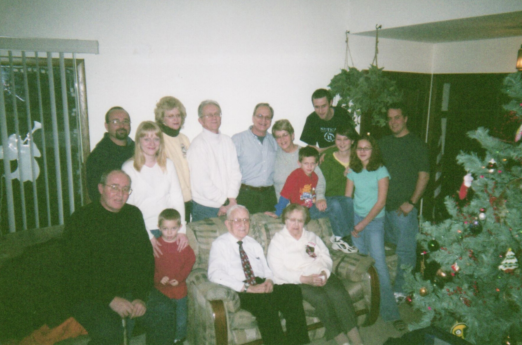 Group of family members of various ages posing for a Christmas photo in a living room with a decorated tree on the right side.
