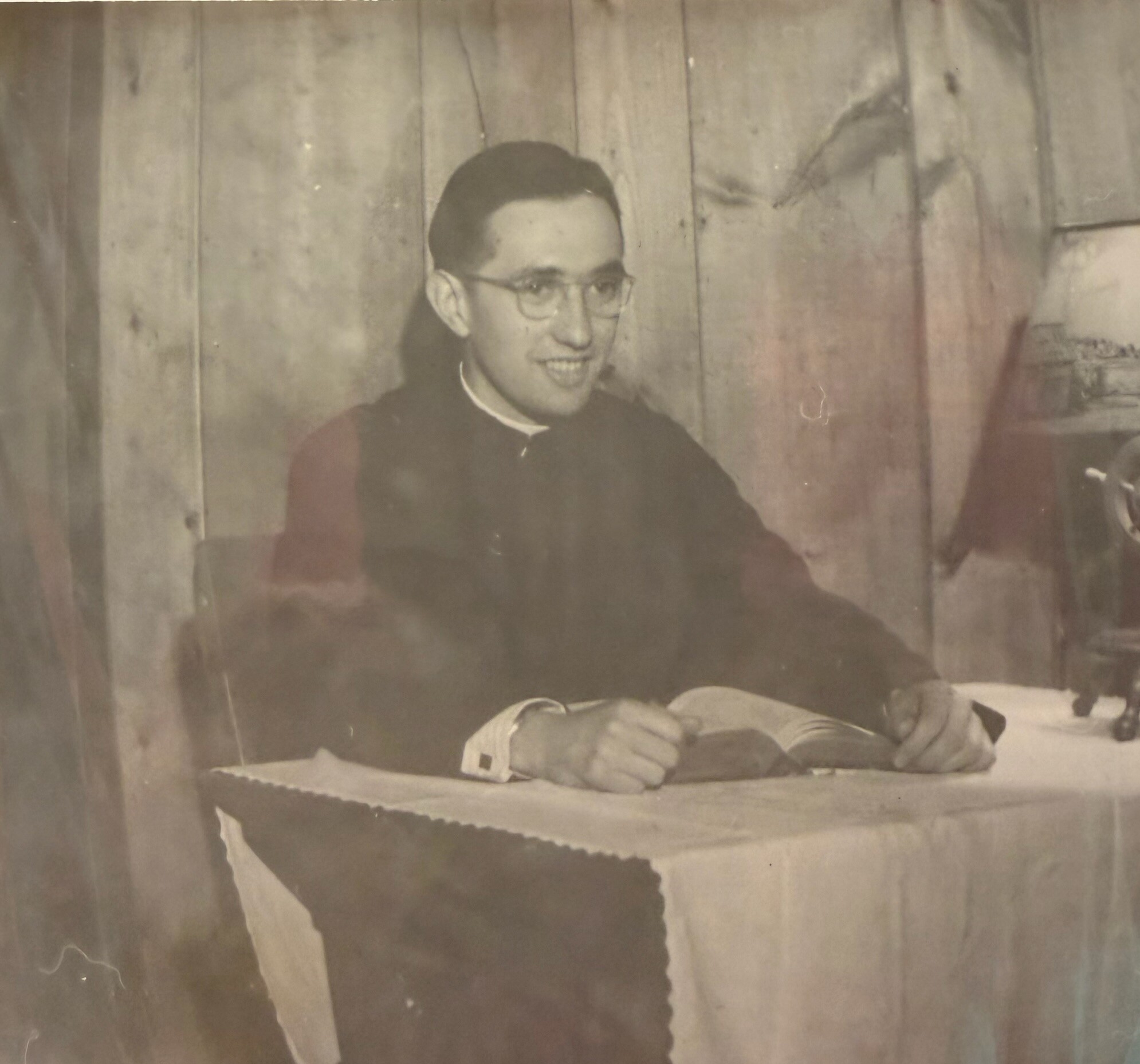 A smiling man with glasses sits at a desk, looking to the left, in a faded vintage photo vignette.