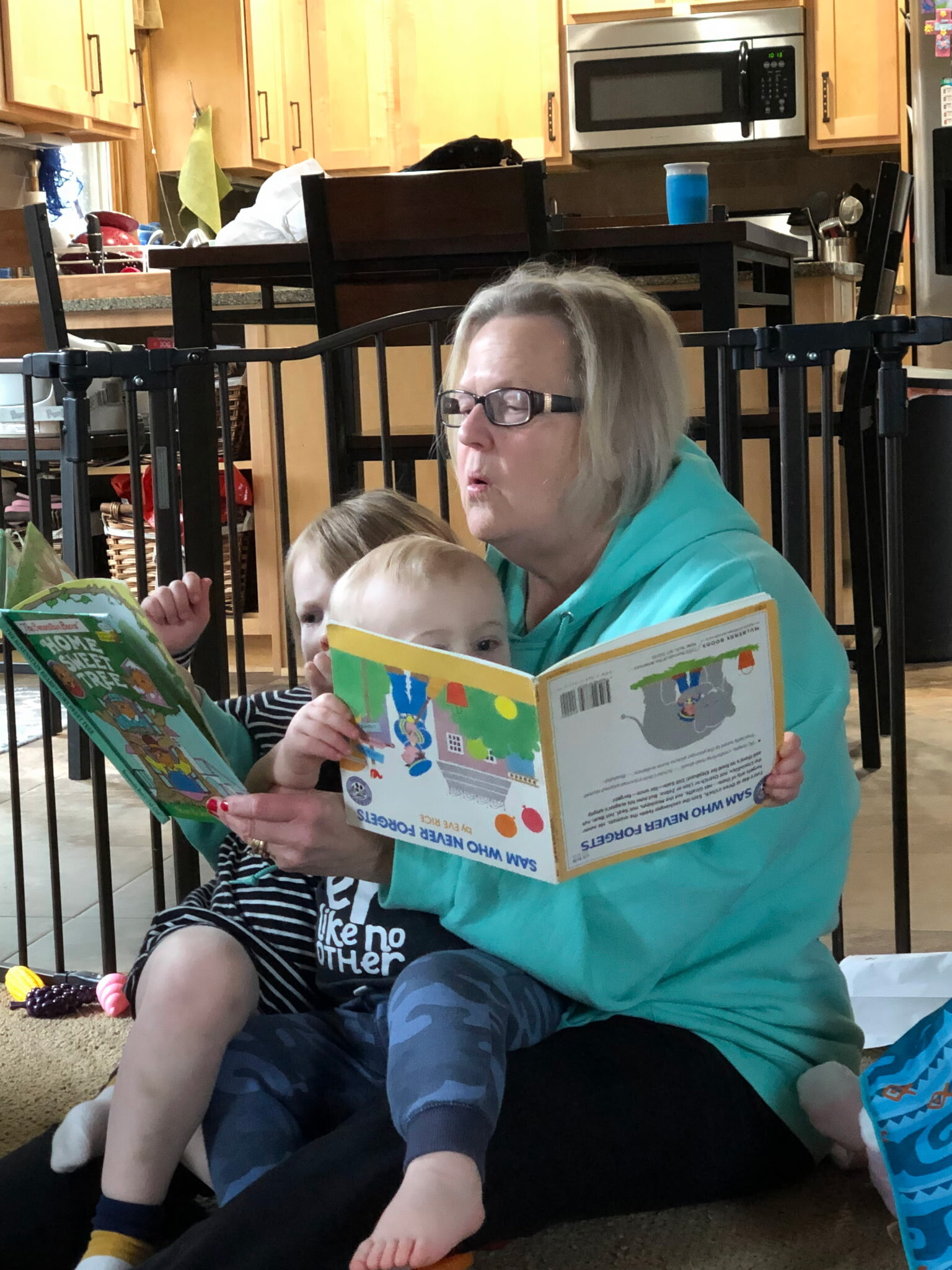 Woman with glasses reads a colorful kids' book to two children sitting on the floor behind a safety gate in a home kitchen.