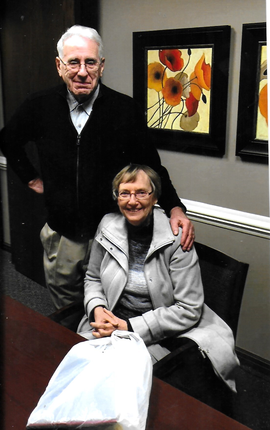 Older man stands behind a seated woman, hand on her shoulder, in a hallway with framed floral wall art.