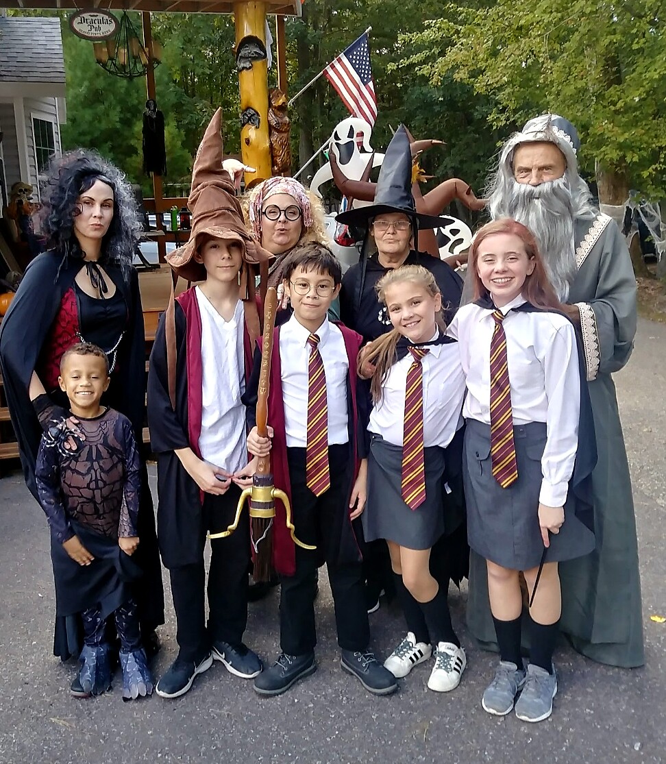 Group of children and adults in Halloween costumes (witches, wizards, students) posing outdoors for a photo, some with wands and a broomstick, smiling.