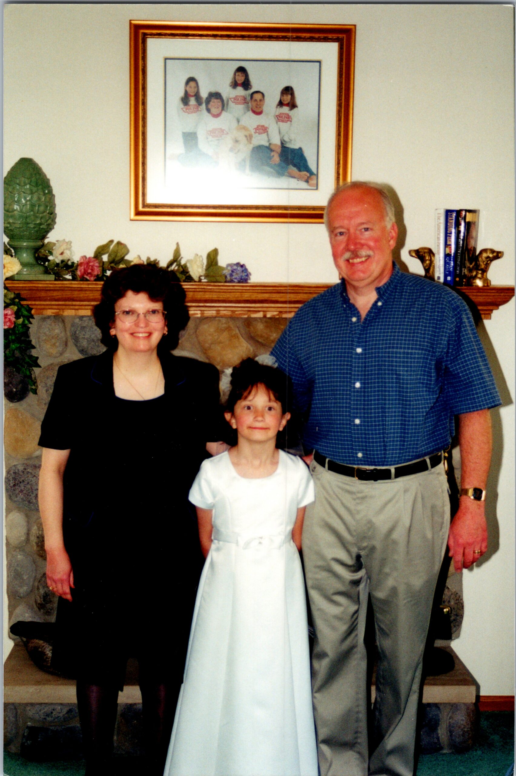 Family posing indoors in front of a stone fireplace, with a framed photo above the mantel: woman in black, a young girl in a white dress, and a man in a blue shirt smile at the camera.