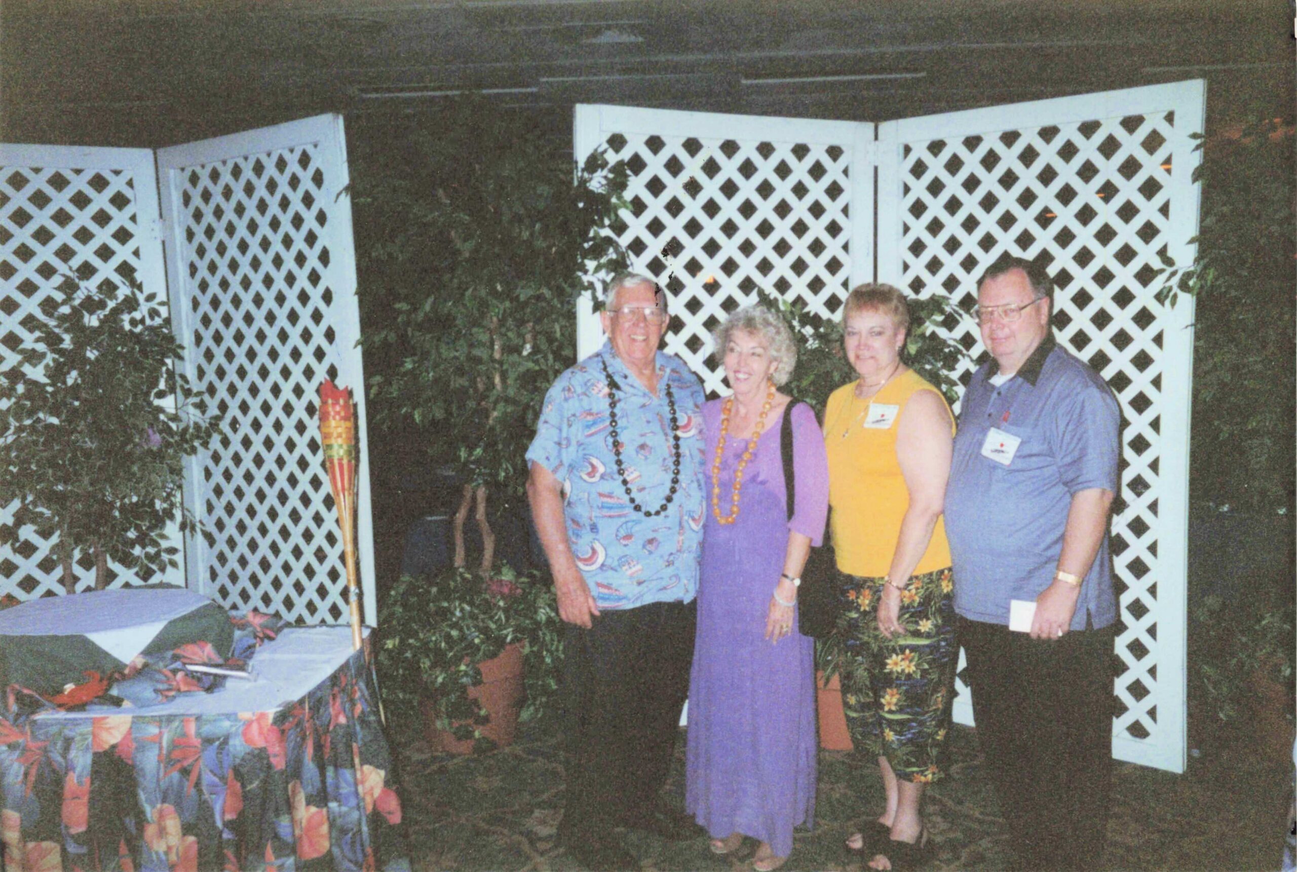 Four adults stand together at a party, wearing colorful outfits and beaded leis, in front of white lattice panels.