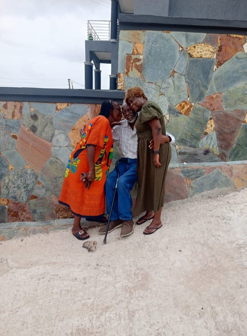 Three people pose together outdoors against a colorful stone wall: an elderly man with a cane sits between two women who lean in for a hug, smiling warmly.
