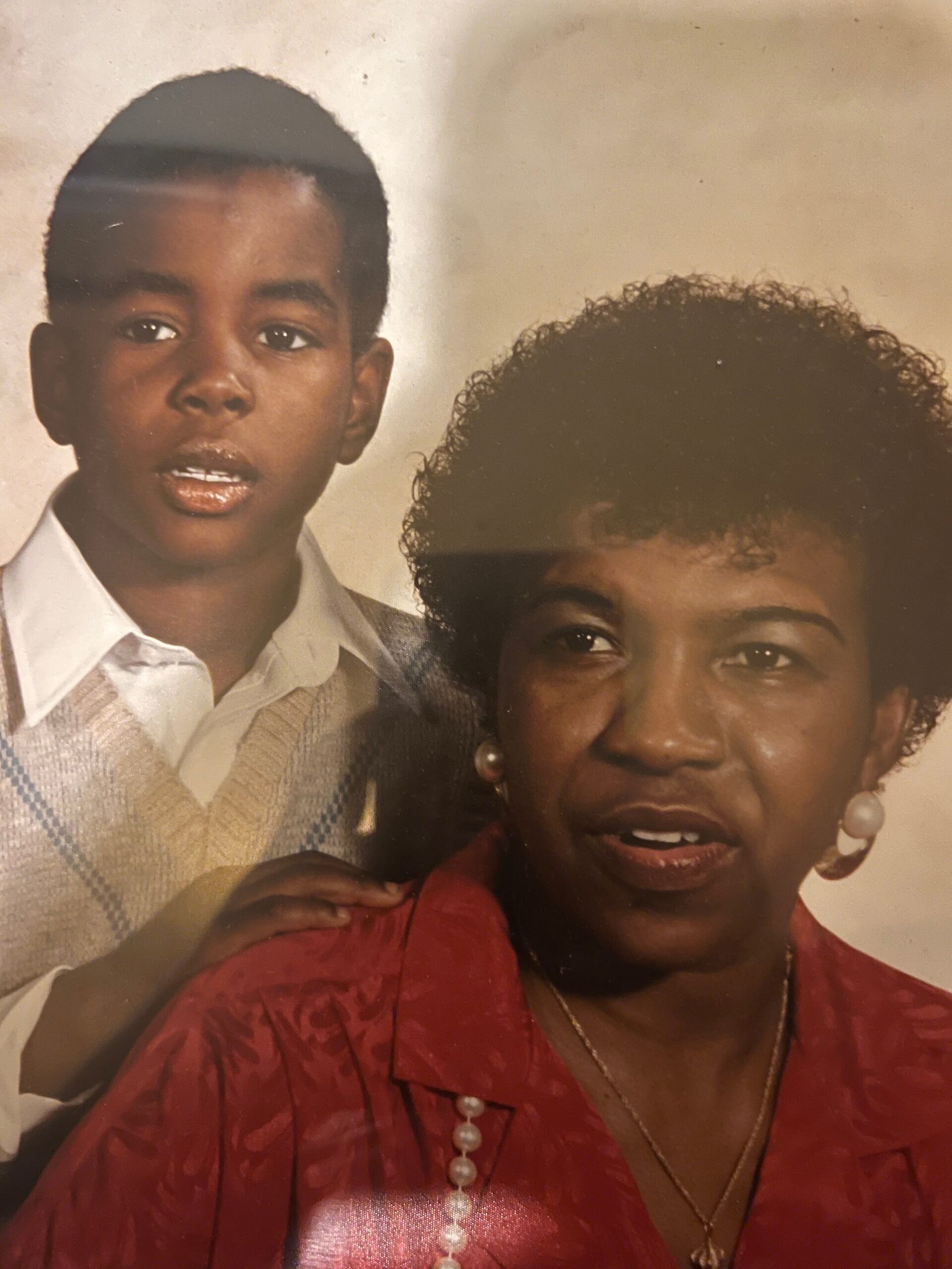 Young boy in a light sweater beside an adult woman in a red blouse and pearl necklace in a studio portrait
