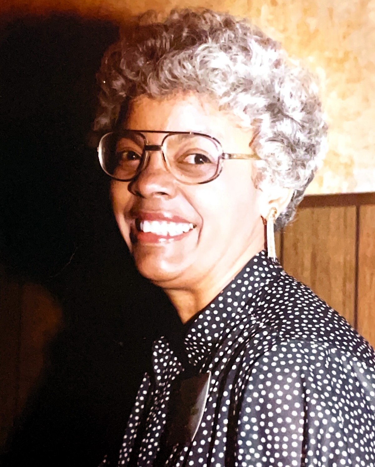 Smiling older woman with short gray hair and glasses, wearing a black polka-dot blouse indoors.