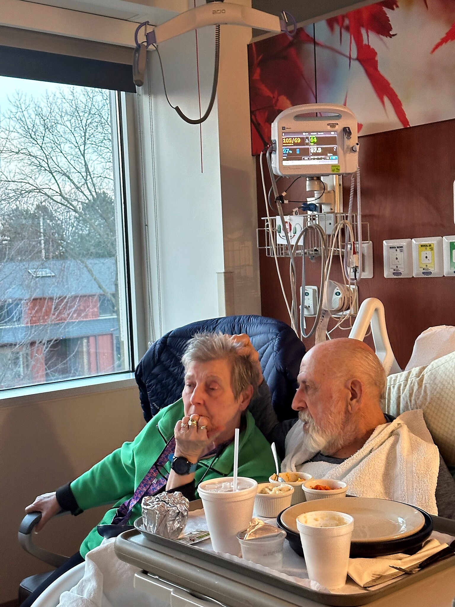 Elderly couple eating at a hospital bedside with a meal tray and drinks, medical monitor and tubing visible in the background, by a window.