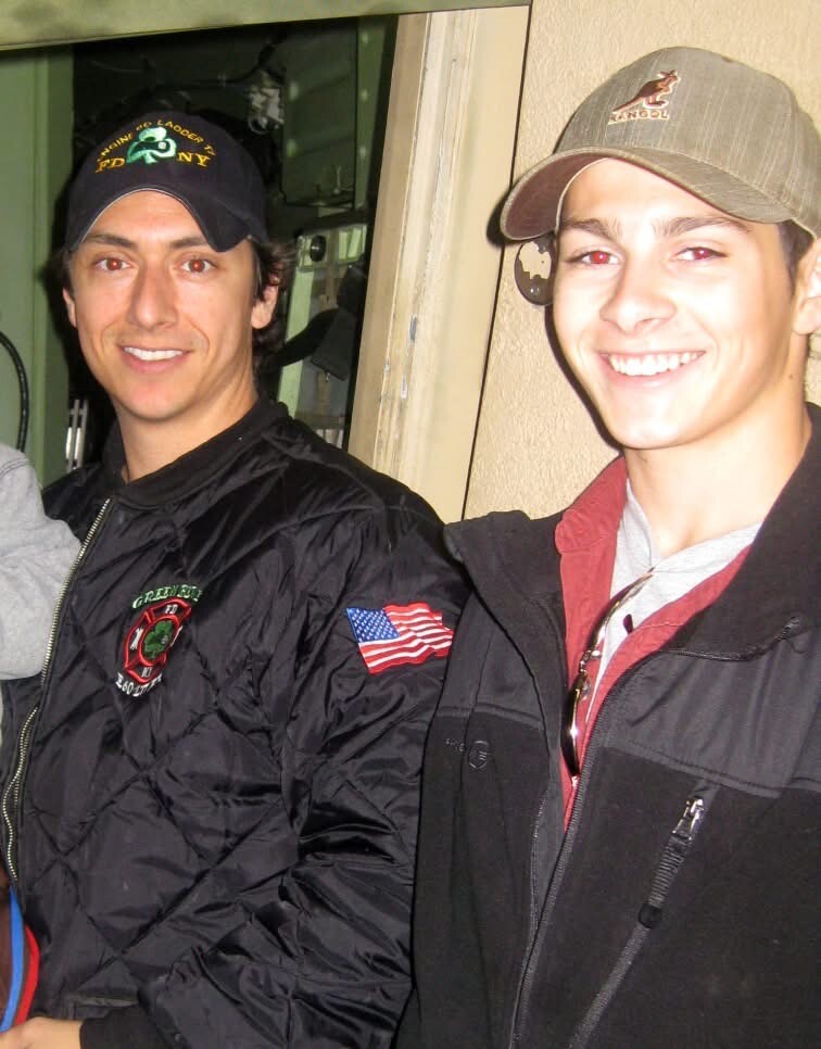 Two young men posing together, one wearing a black jacket with an American flag patch on the sleeve and a dark cap, smiling at the camera.
