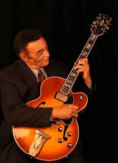 A man in a suit playing an orange sunburst jazz guitar against a dark background.