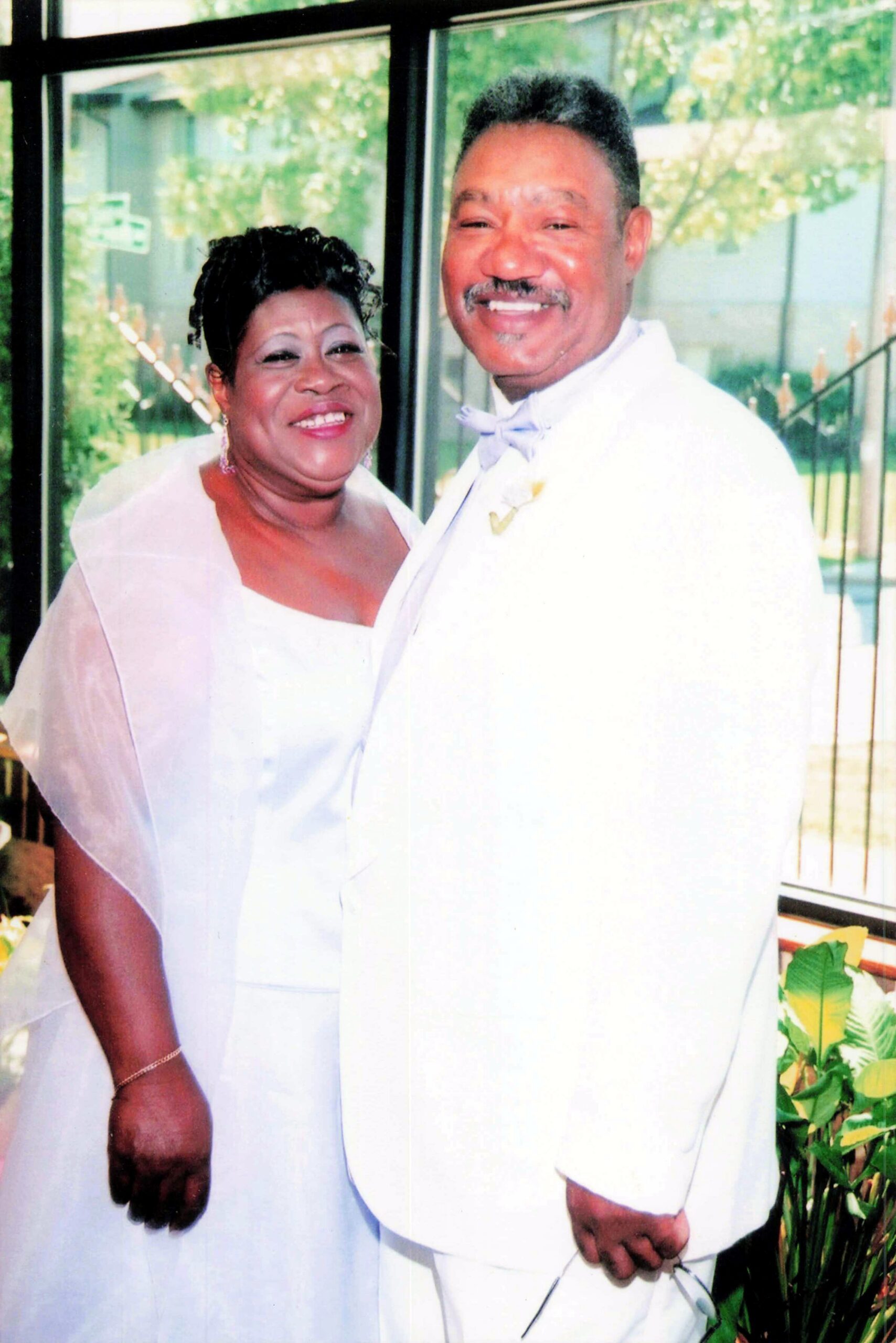 Smiling African American couple in wedding attire pose together indoors with a glass wall and greenery in the background.
