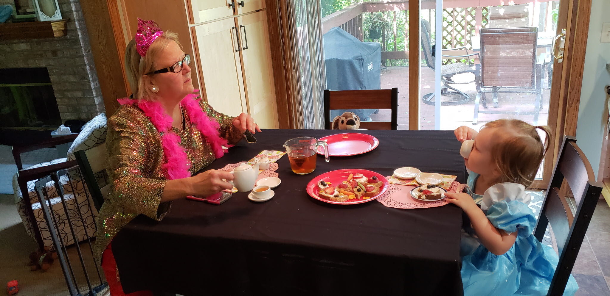 Older woman in a glittery gold jacket with a pink tiara and boa sits at a dining table with a young girl in a blue princess dress, enjoying tea and treats.