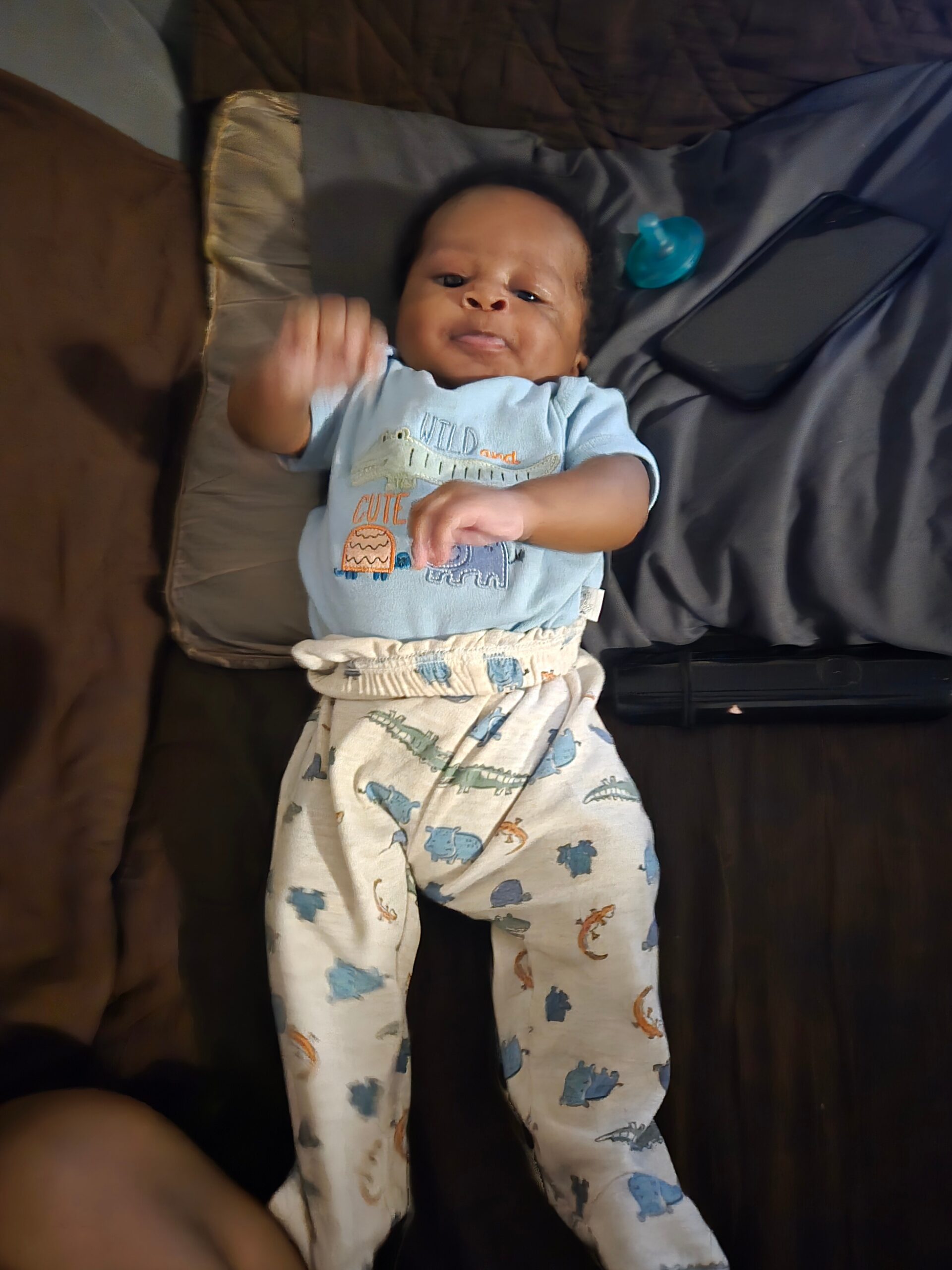 Baby lying on a bed in a light blue outfit with animal prints; a blue pacifier rests on a gray pillow nearby.