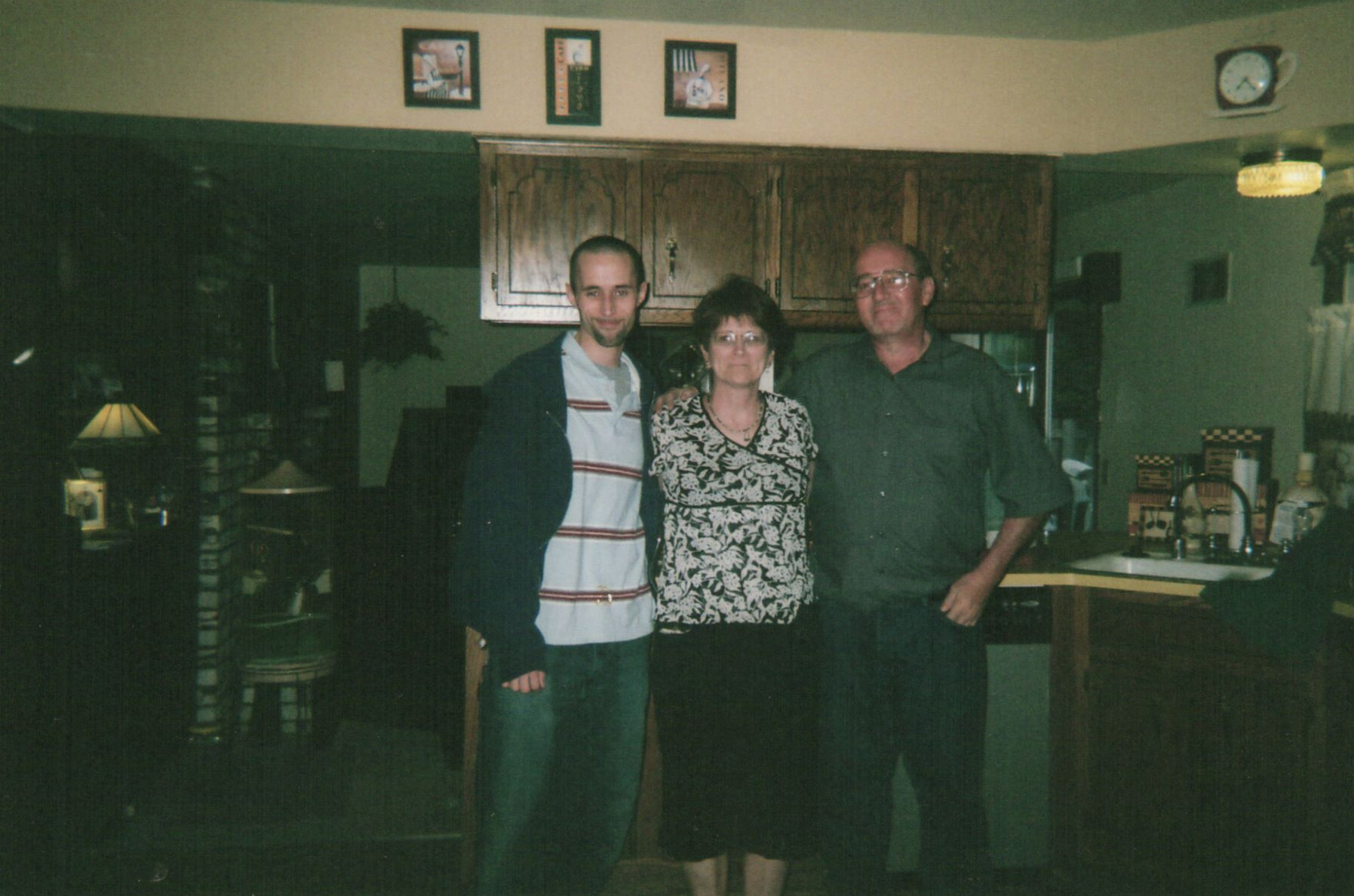 Three adults pose together in a home kitchen, standing side by side in front of wooden cabinets.