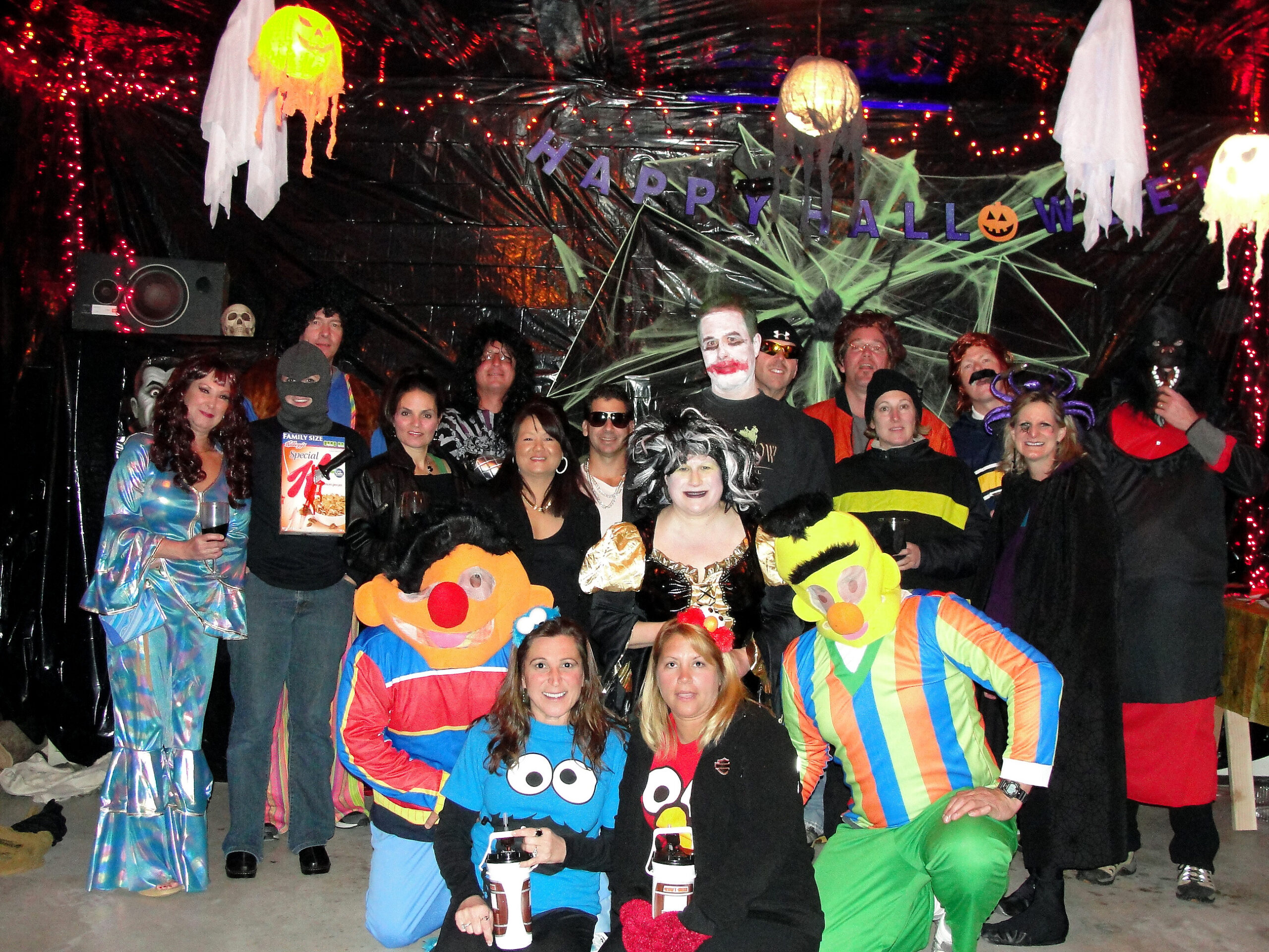 Group of adults in colorful Halloween costumes posing for a photo, with a 'Happy Halloween' banner and cobweb decorations in the background.