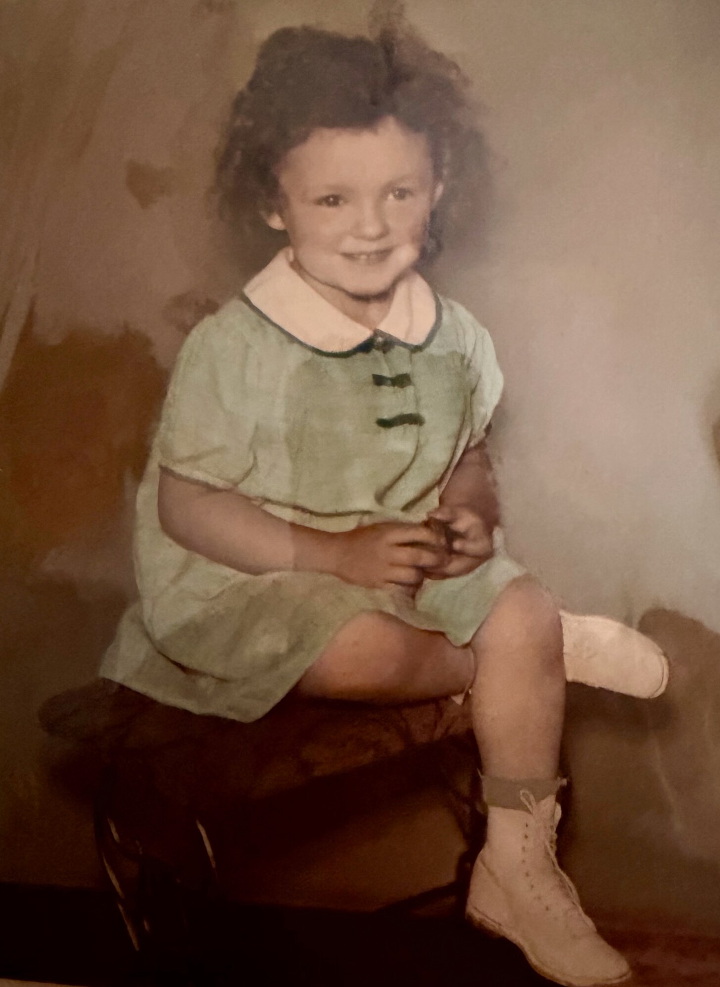 Young child with dark hair, wearing a pale green dress and white collar, sitting and smiling while holding a small object; left leg in a cast or bandage.