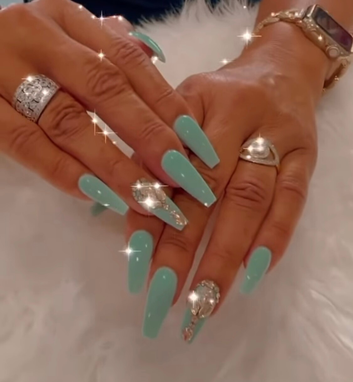 Hands with long turquoise coffin nails, wearing crystal rings and a gold bracelet. Magnified manicure close-up.