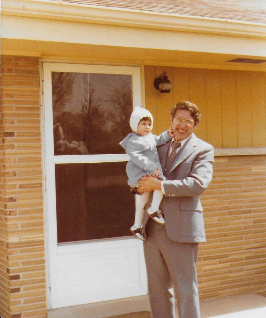 Man in a gray suit holding a baby in a light outfit, standing in front of a brick house with a white door.