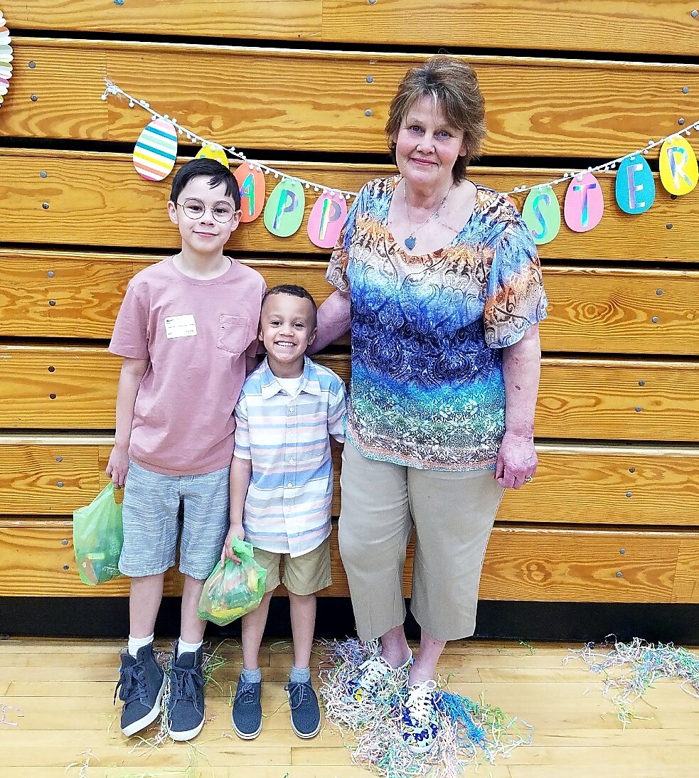 Grandmother with two boys posing at an Easter celebration, colorful banner in the background and confetti on the floor.