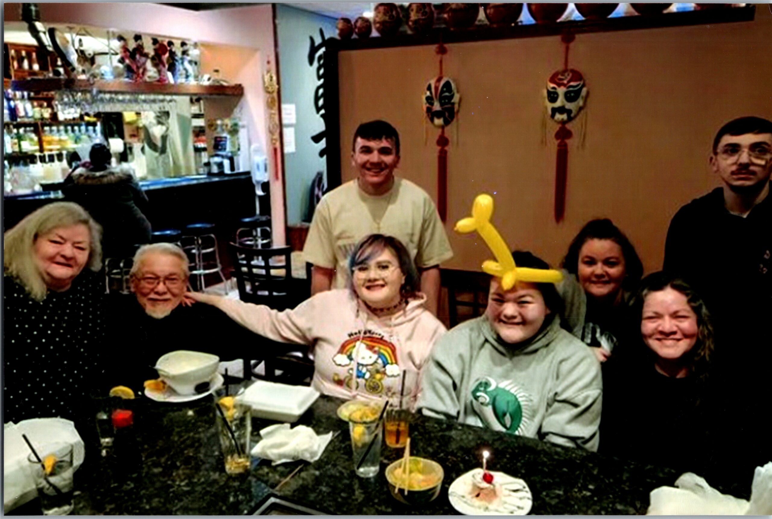 Group of smiling people around a restaurant table celebrating a birthday, with cake, balloons, and party hats visible in the background.