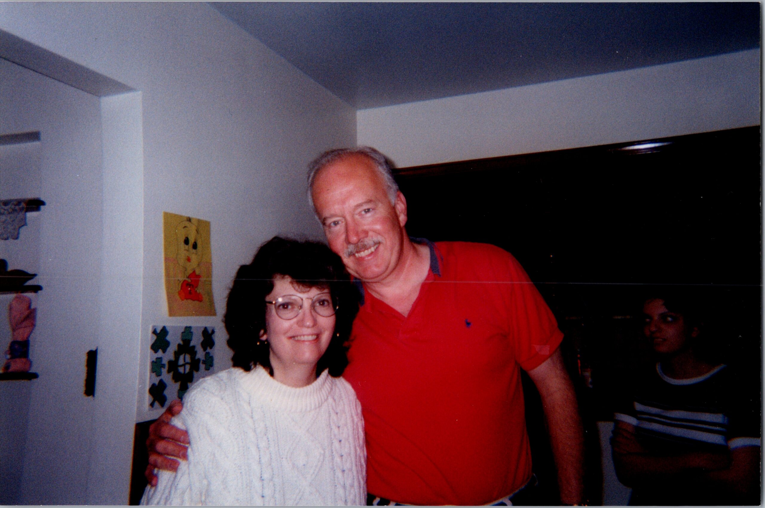 Smiling man in a red shirt stands with a woman in a white knit sweater indoors, arm around her shoulders.