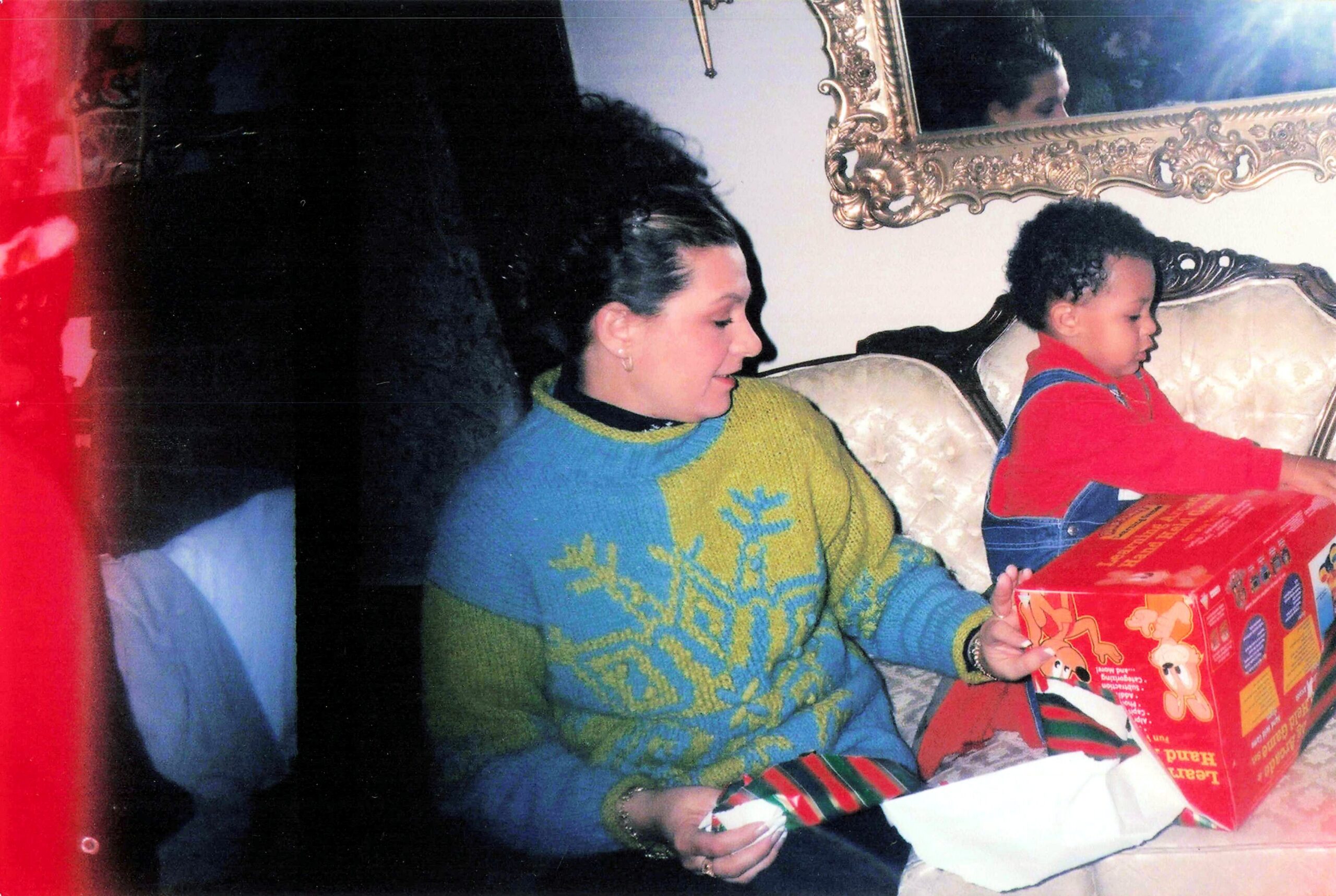 Woman in a blue and yellow sweater sits next to a child in red overalls, both looking at a large red striped gift box on an ornate couch.