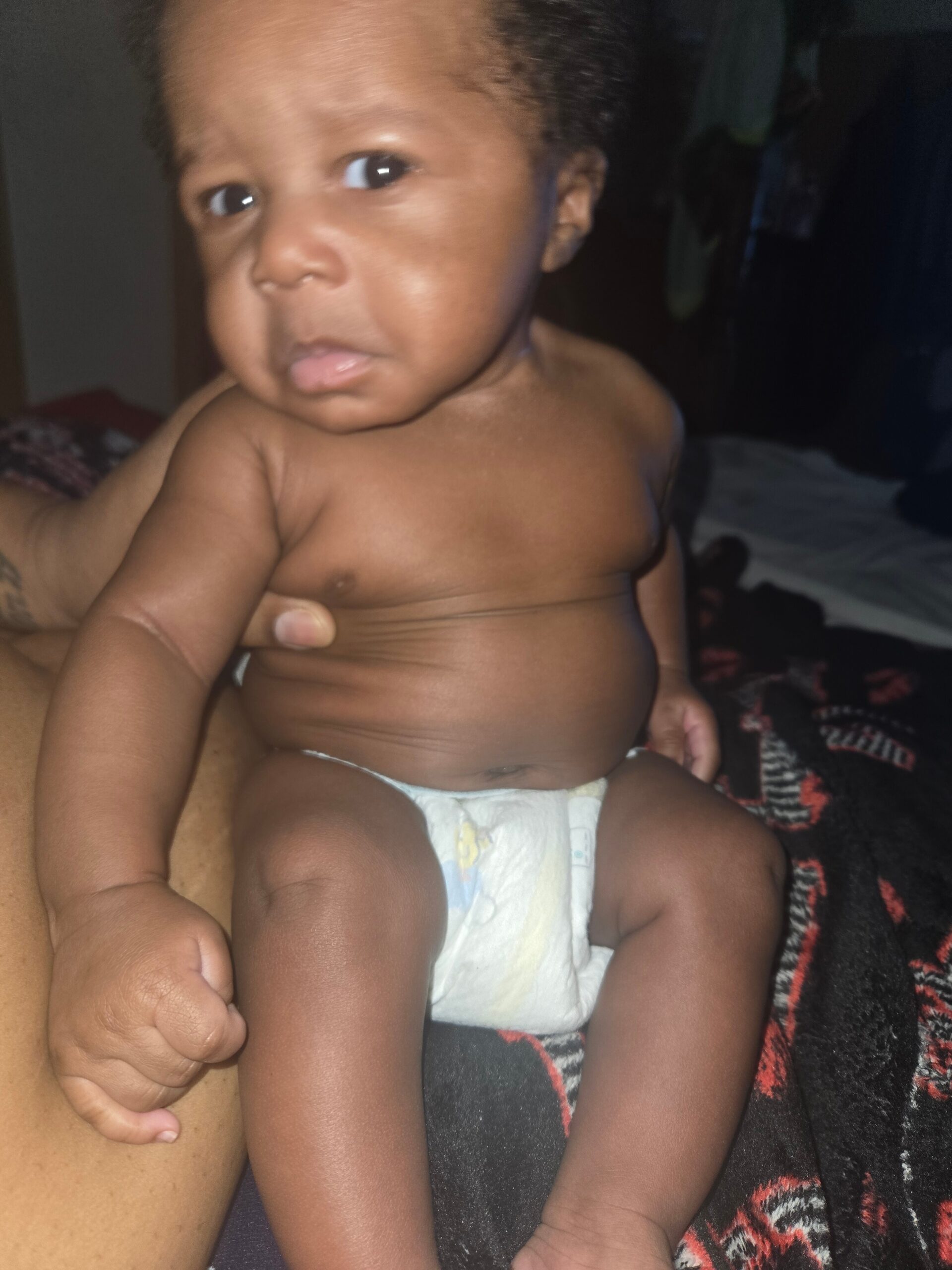 Close-up of a baby being held by an adult, shirtless and wearing a diaper, looking at the camera in a dim room.