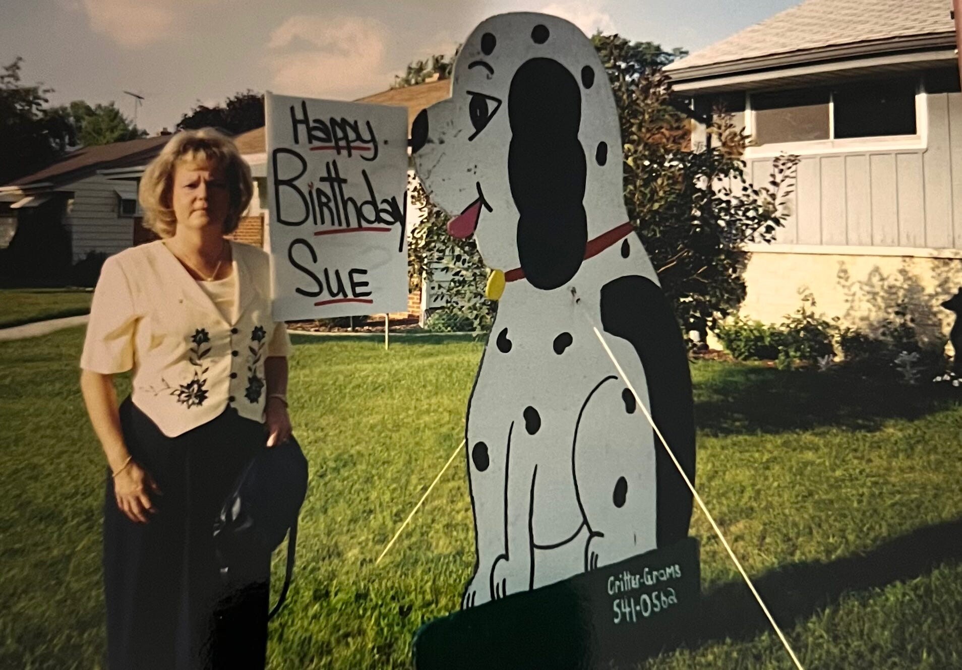 Woman posing next to a large cartoon snowman cutout and a 'Happy Birthday Sue' sign in a suburban front yard.
