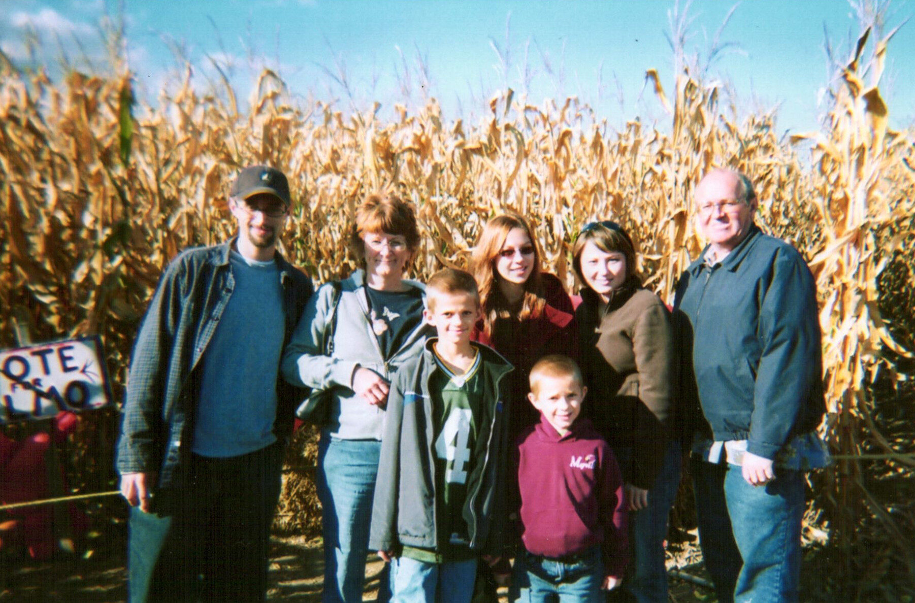 Group of multiple family members posing together in a cornfield on a sunny day, smiling at the camera before tall dried cornstalks.