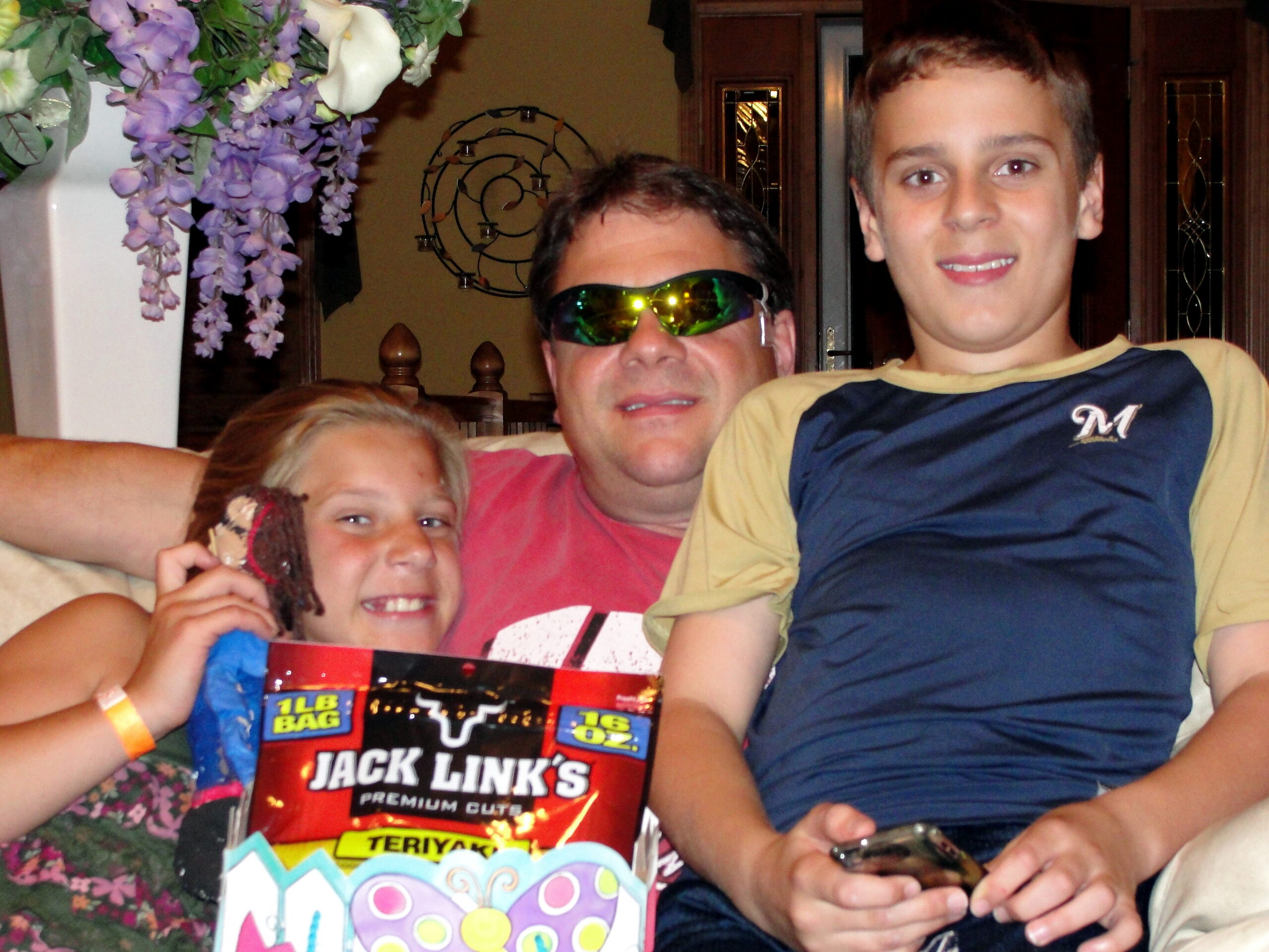 Three family members pose for a casual party photo: a man wearing sunglasses in the center, a boy on the right, and a girl on the left, holding a Jack Link's snack package and a colorful gift bag behind them with flowers nearby.